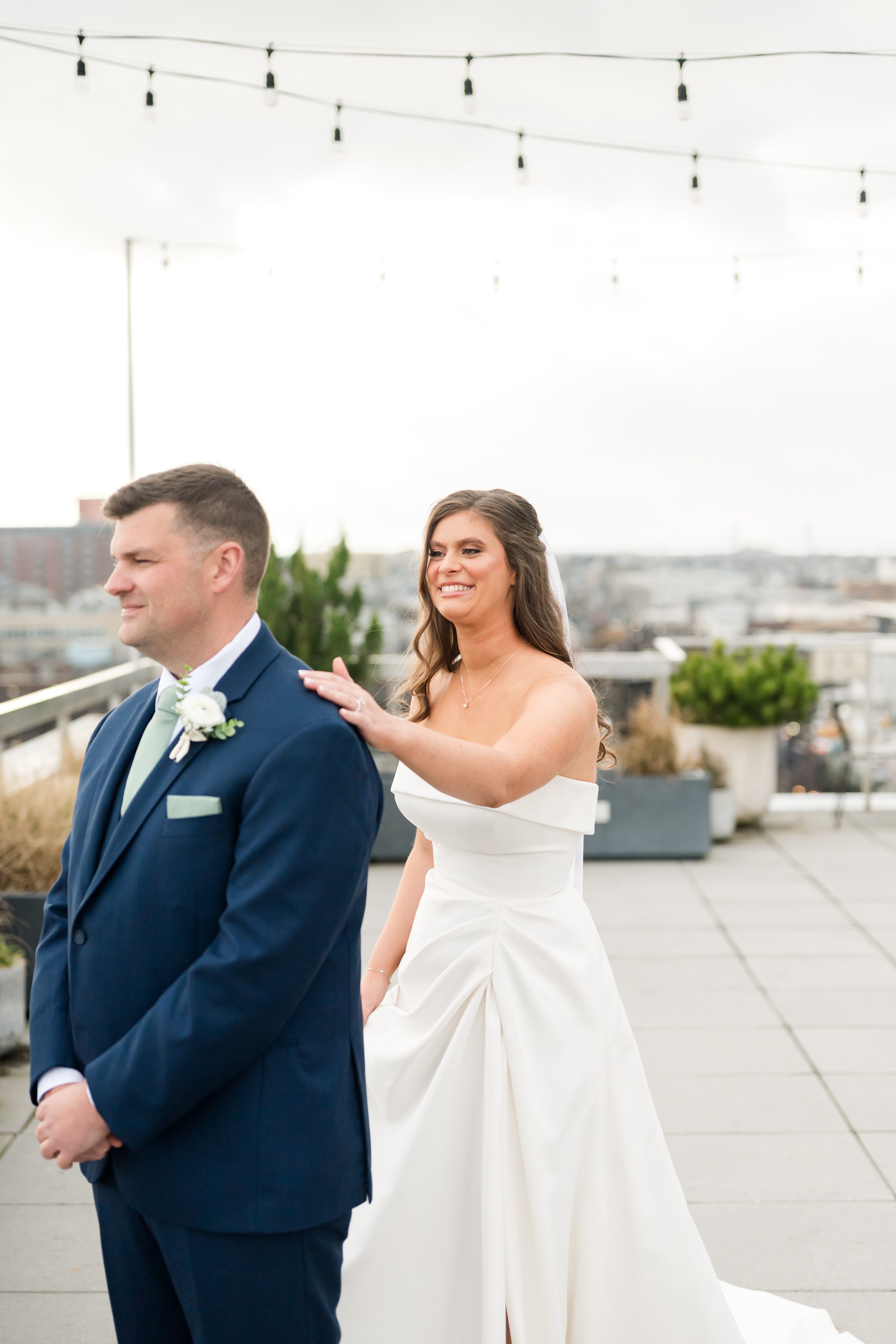 Bride taps groom’s shoulder during first look on a rooftop with string lights overhead.