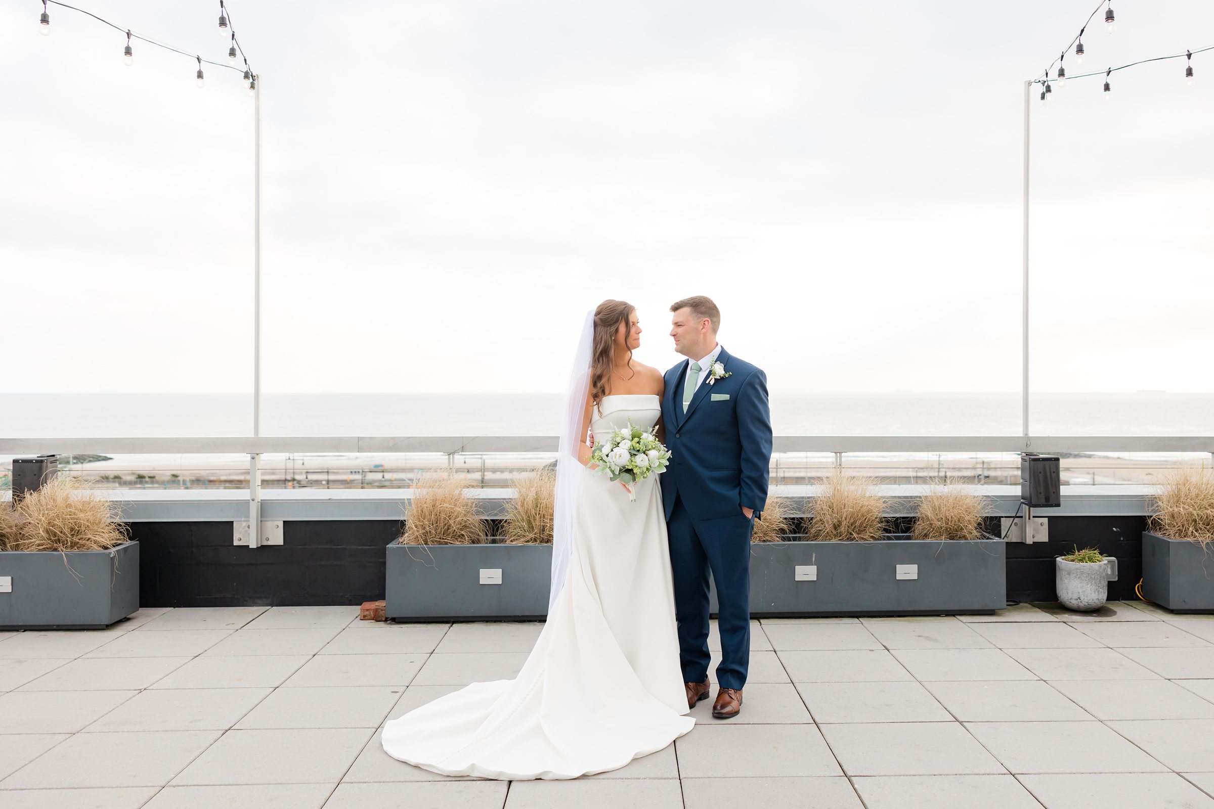 Bride and groom standing together on rooftop with string lights and ocean view.
