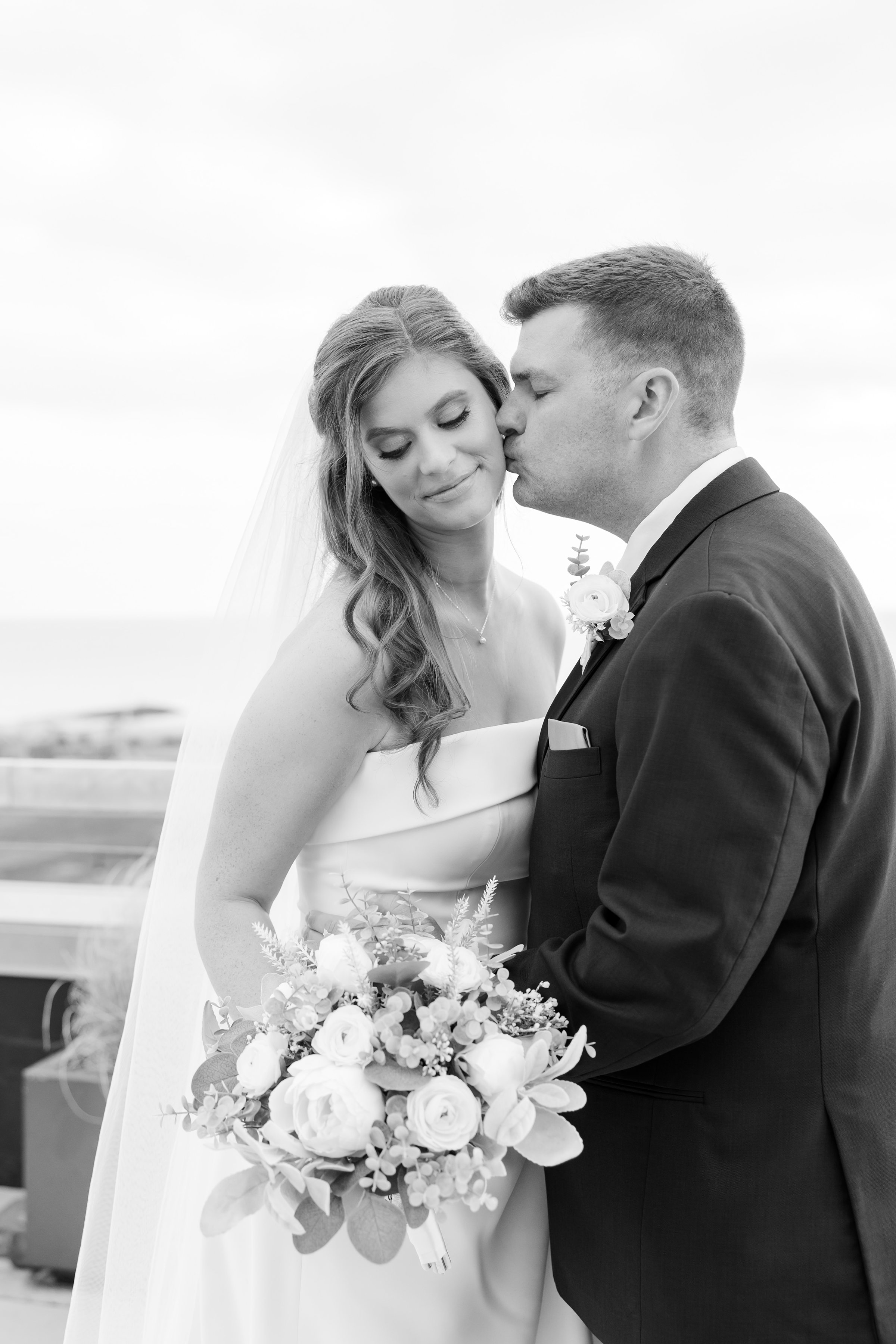 Bride and groom kissing bride’s cheek while she holds bouquet.