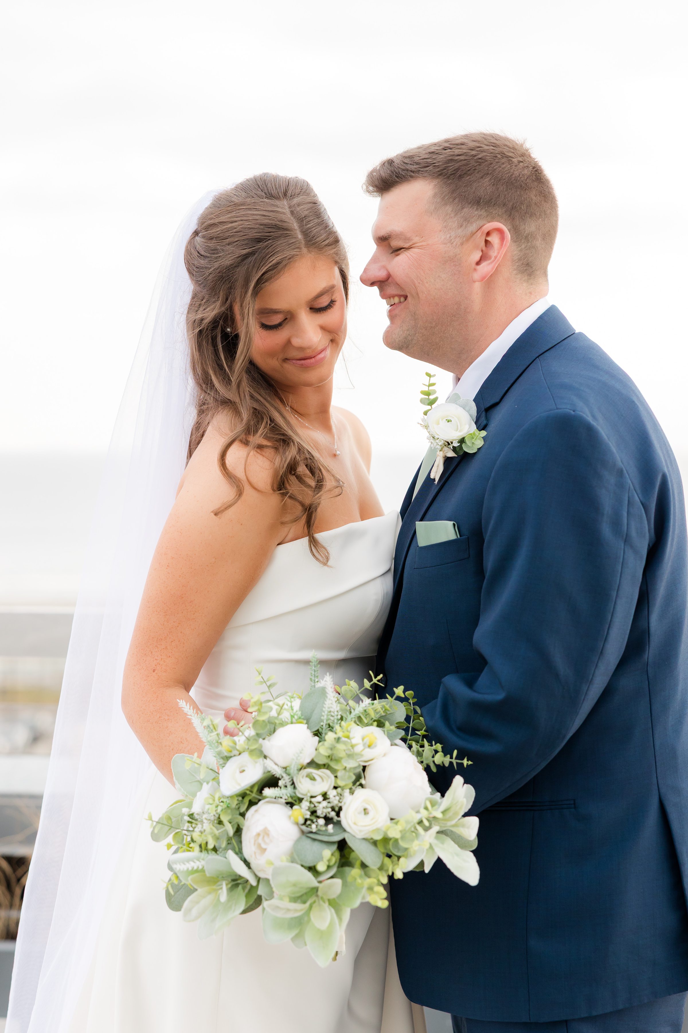 Bride and groom holding bouquet together and smiling on rooftop with ocean in background.