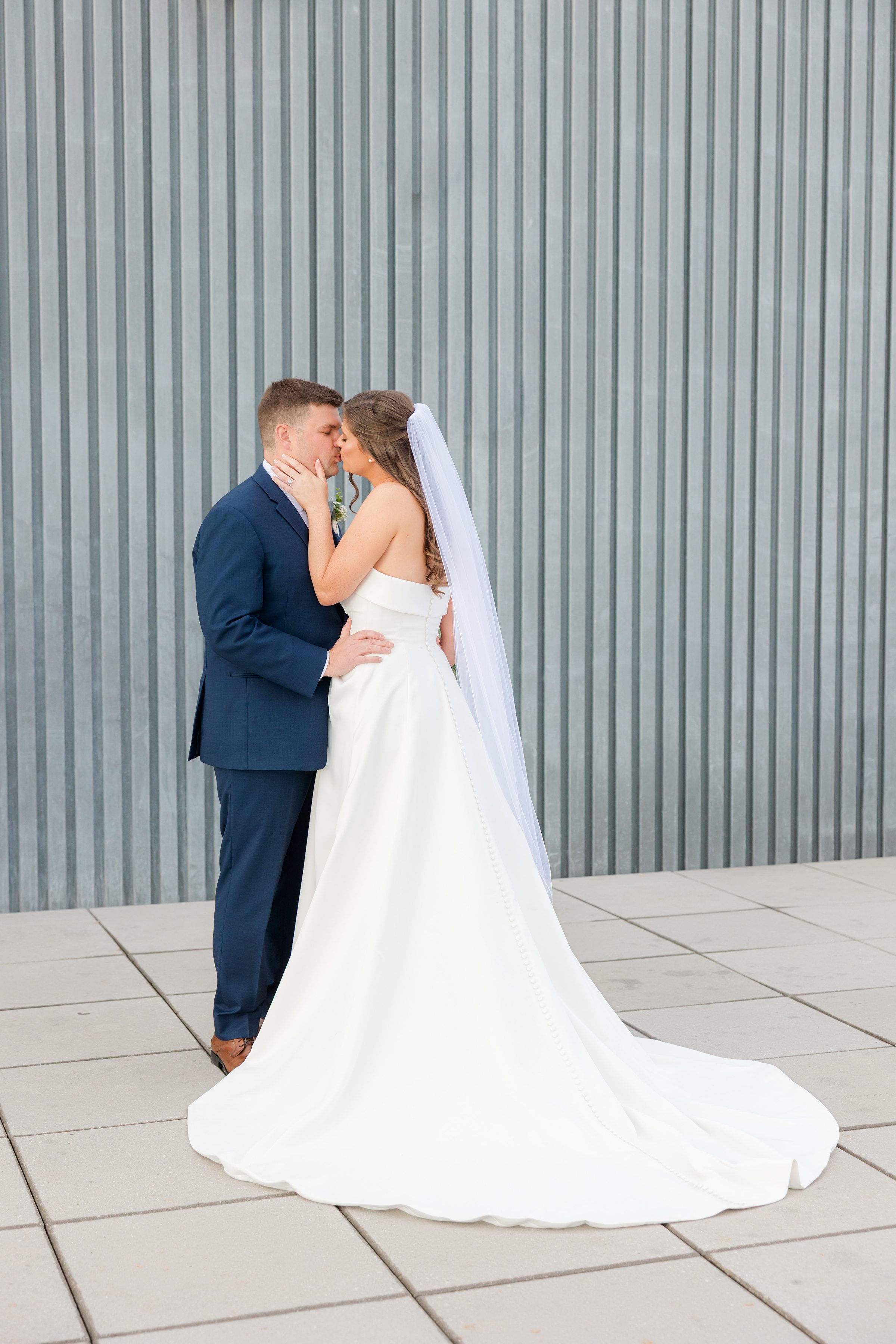 Bride and groom share a tender kiss, holding each other close against a textured wall, her veil softly cascading behind her as they pause in a quiet, intimate moment together.