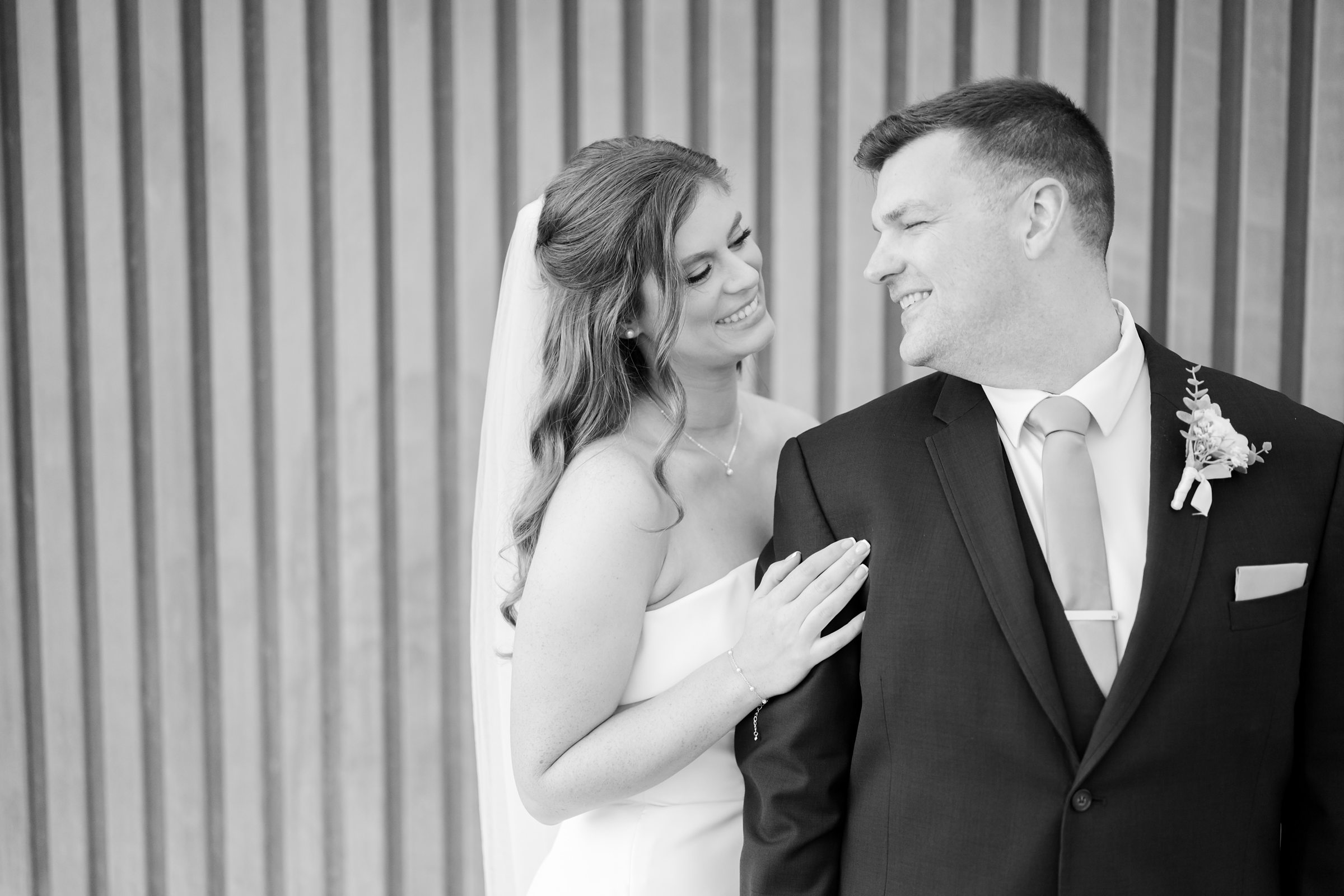 Bride gently resting her hand on the groom’s shoulder as they smile at each other, sharing a soft, affectionate moment filled with warmth and love.
