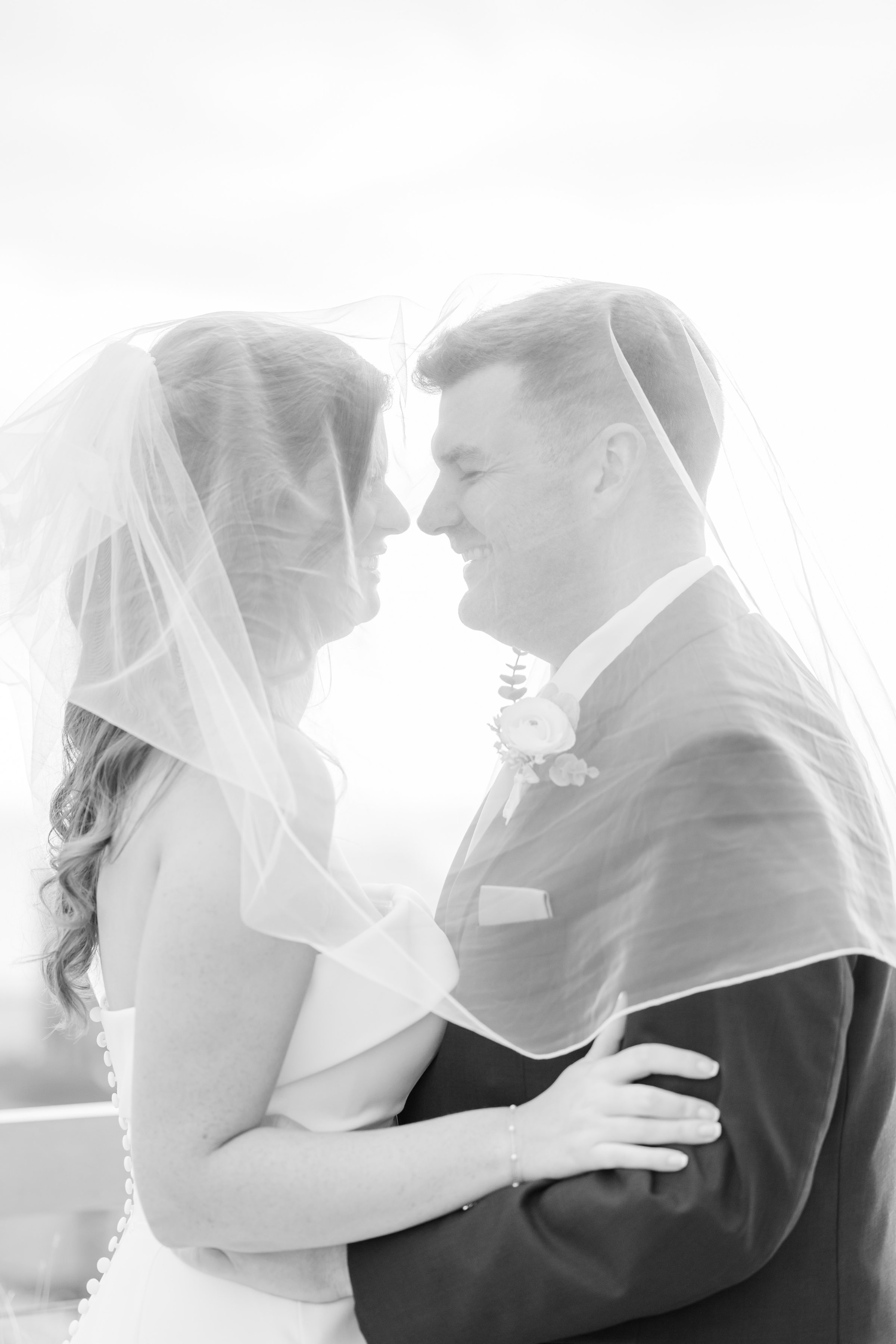 Bride and groom smiling beneath her veil, their faces close as they share a tender, intimate moment wrapped in light and love.