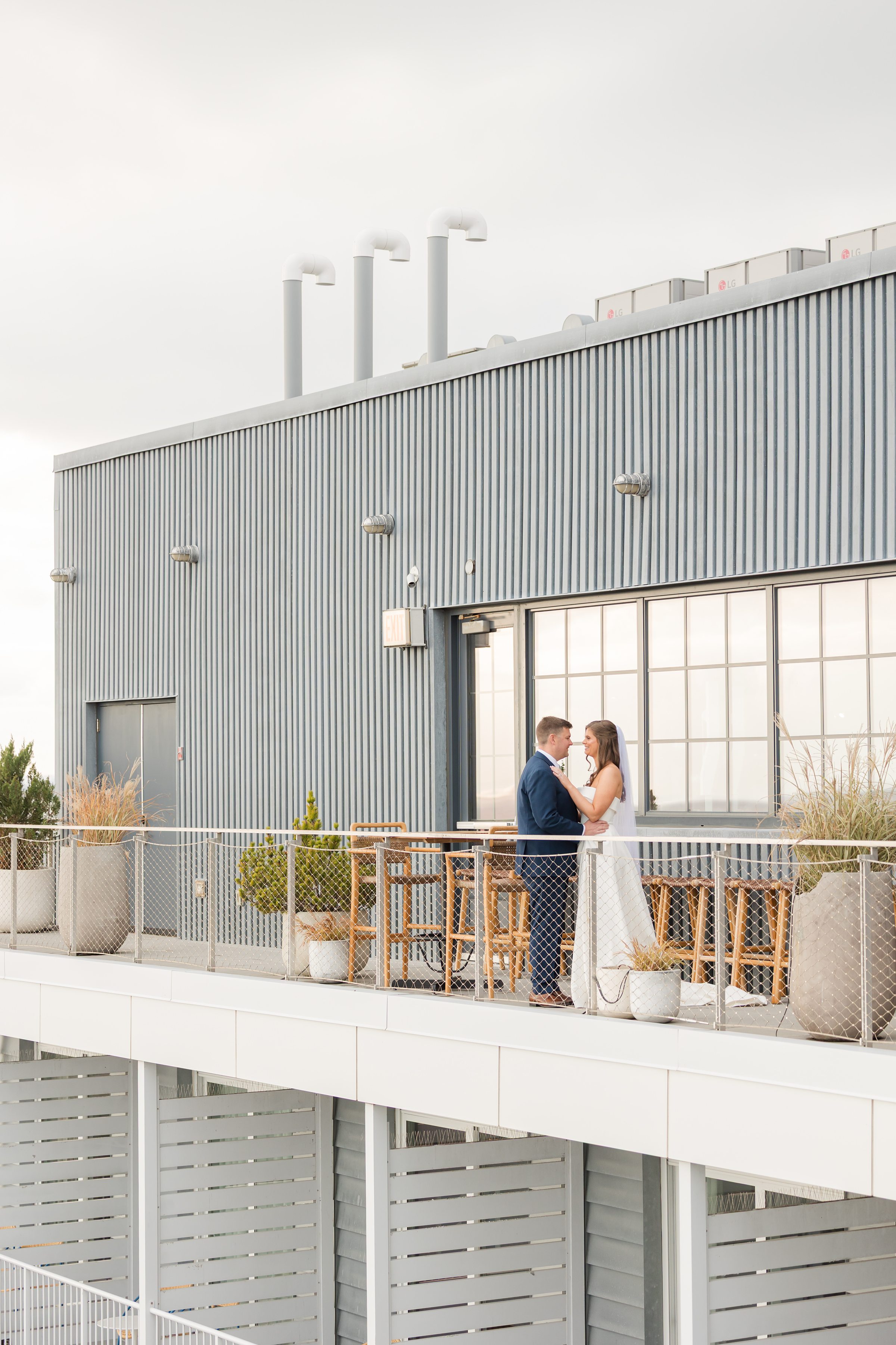 Bride and groom embracing on a modern rooftop balcony with metal siding and potted plants.