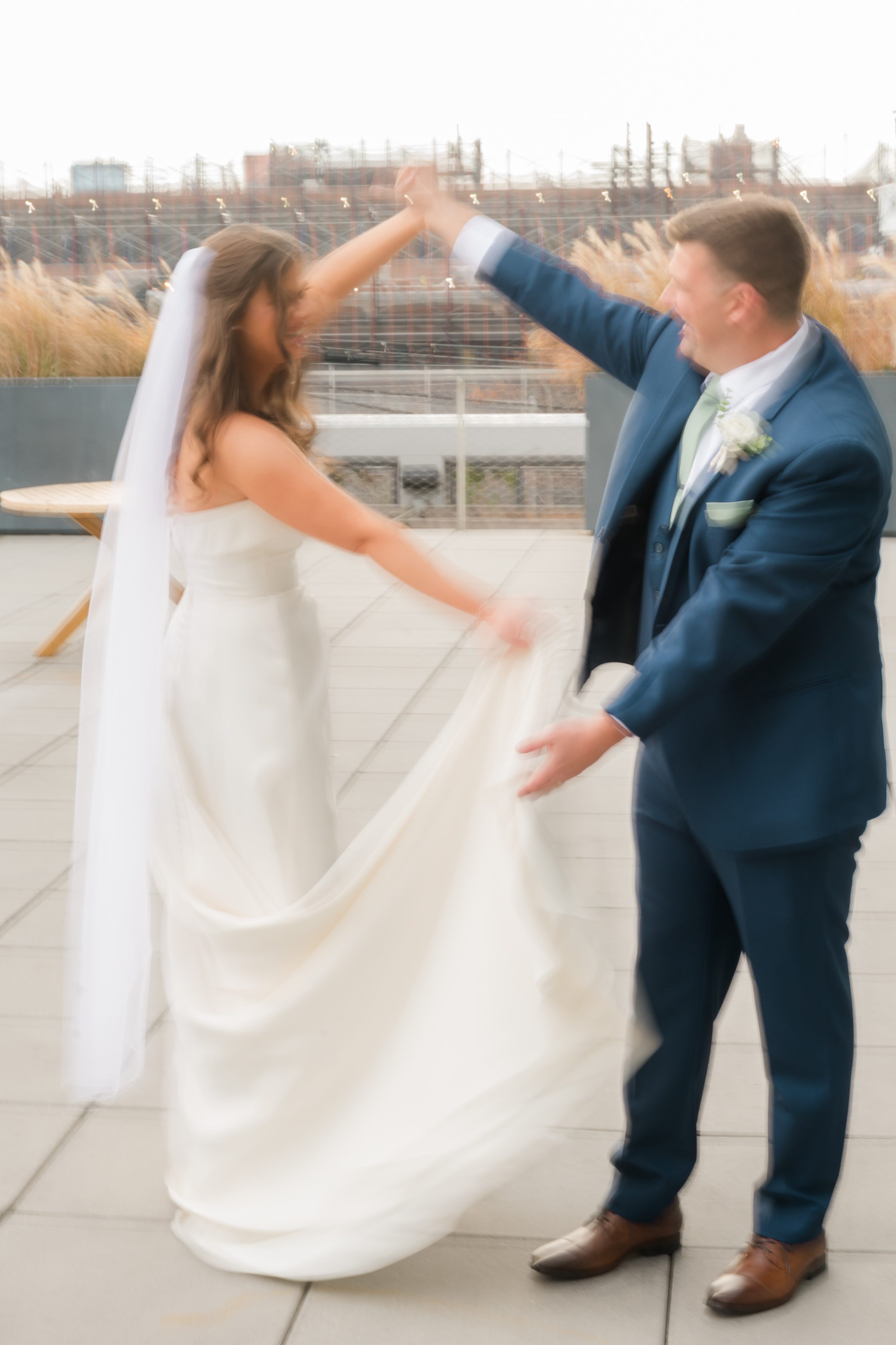 Bride and groom share a joyful, romantic moment as he twirls her beneath soft golden light, her veil and dress flowing gracefully in the air on a rooftop terrace.