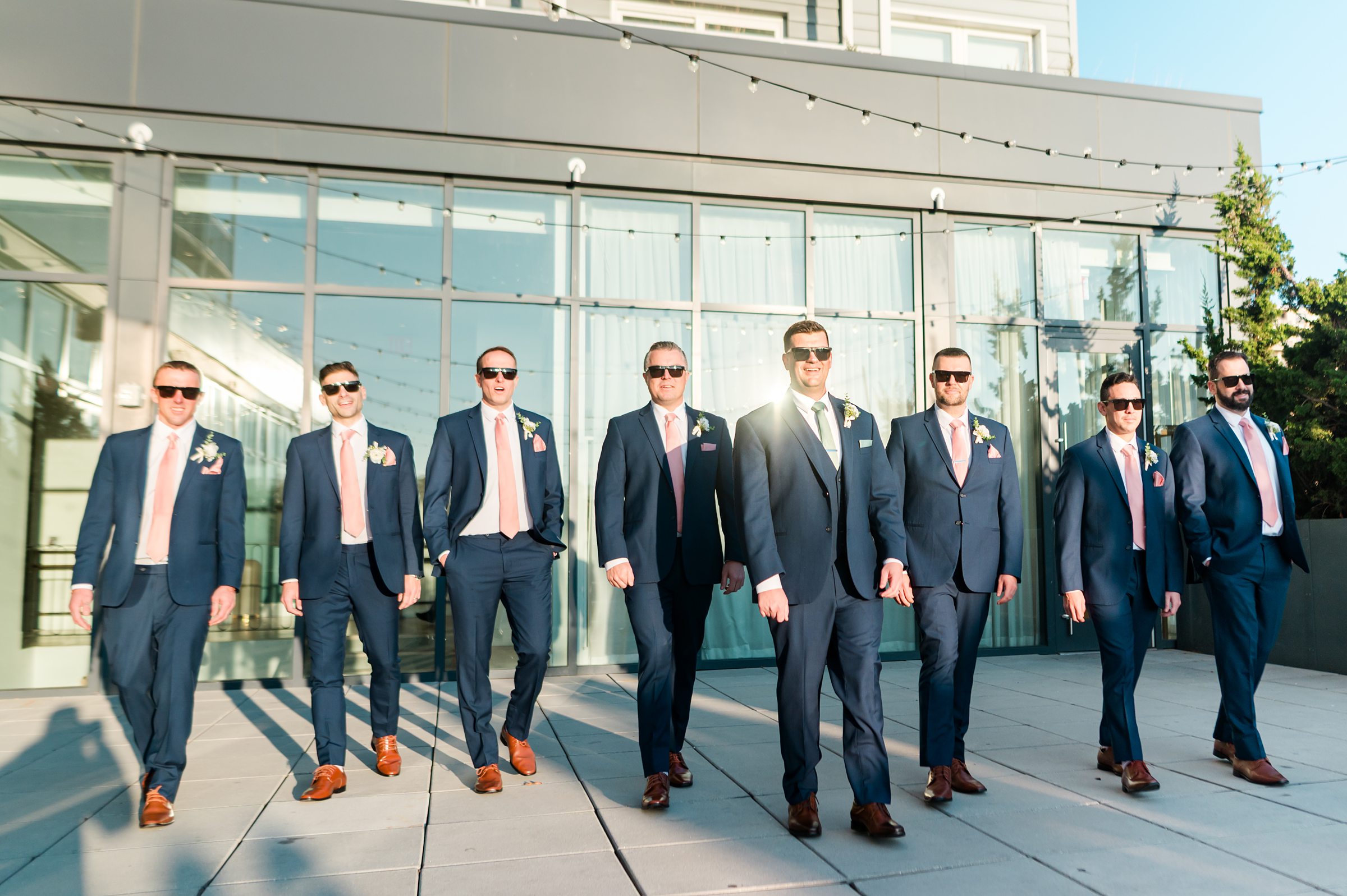 Groom and groomsmen in matching navy suits and sunglasses walk confidently together outdoors in bright sunlight.