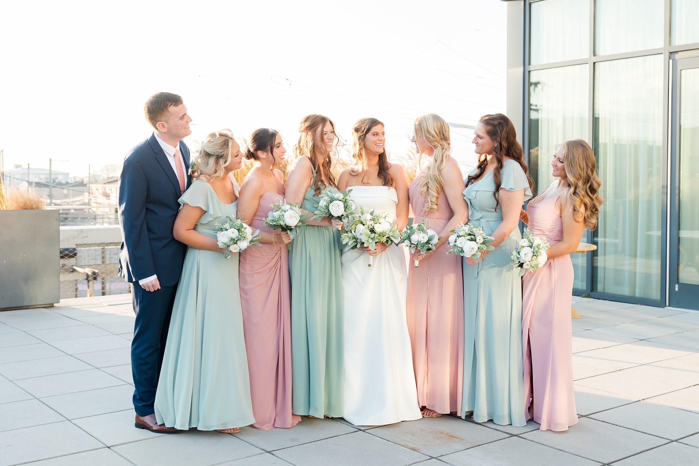 Bride surrounded by bridesmaids in pastel dresses holding bouquets, all smiling together.
