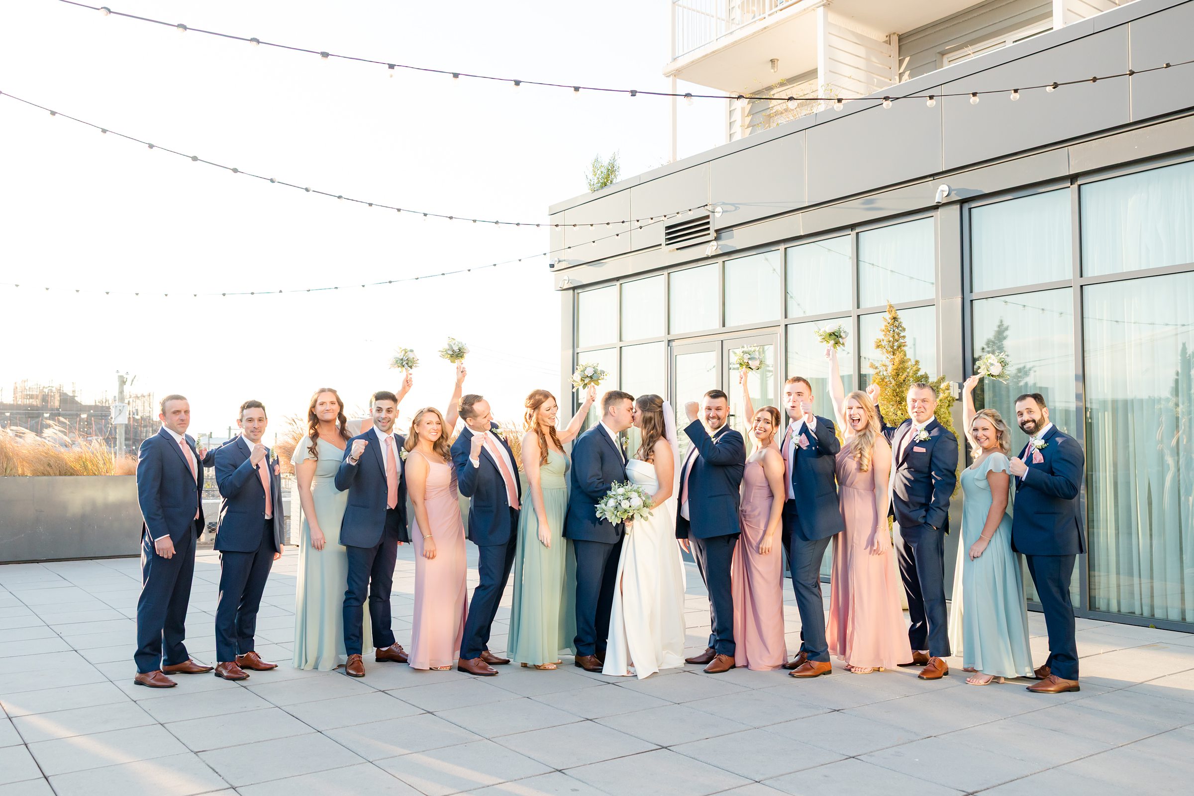 Bride and groom celebrate with their bridesmaids and groomsmen, all smiling and cheering together on a sunlit rooftop.