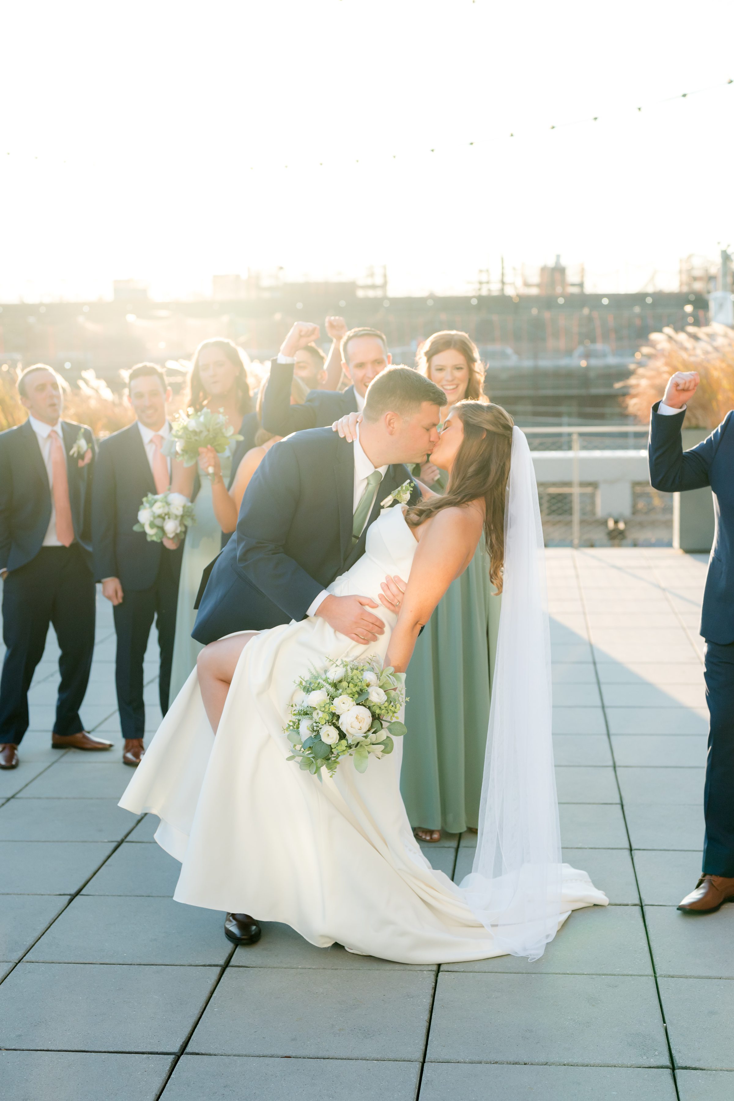 Bride and groom kissing at sunset while wedding party cheers behind them on a rooftop.