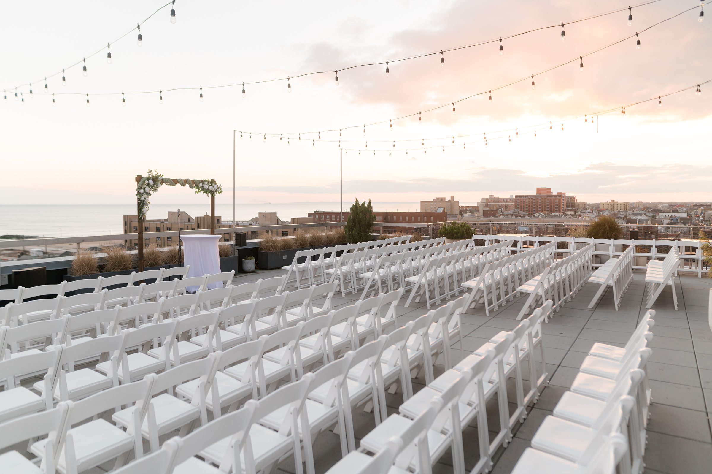 Rooftop wedding ceremony setup with rows of white chairs, a floral arch, and string lights overlooking the ocean at sunset.