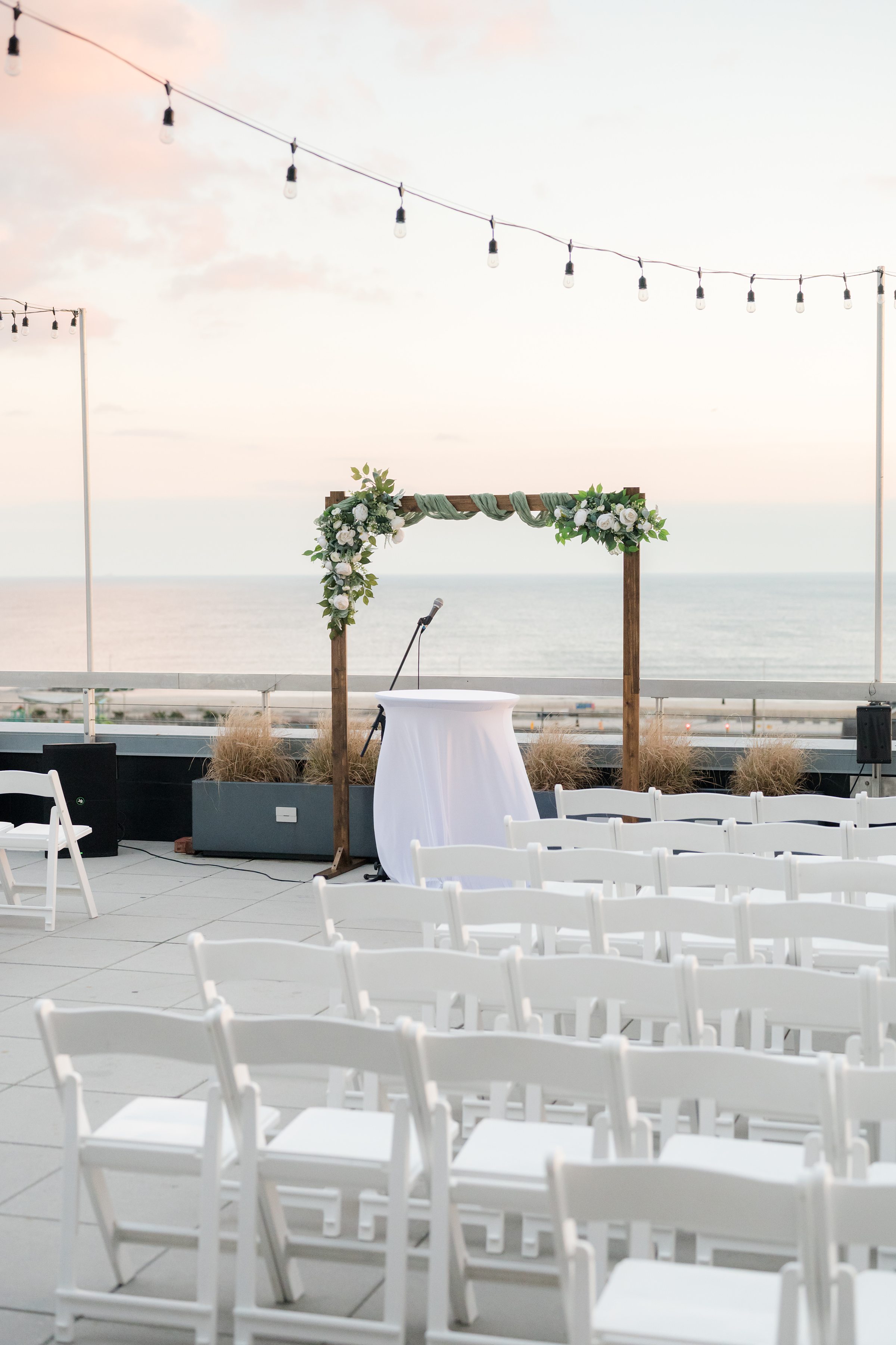 Floral wedding arch and ceremony space set on a rooftop with ocean views, softly lit by string lights at sunset.