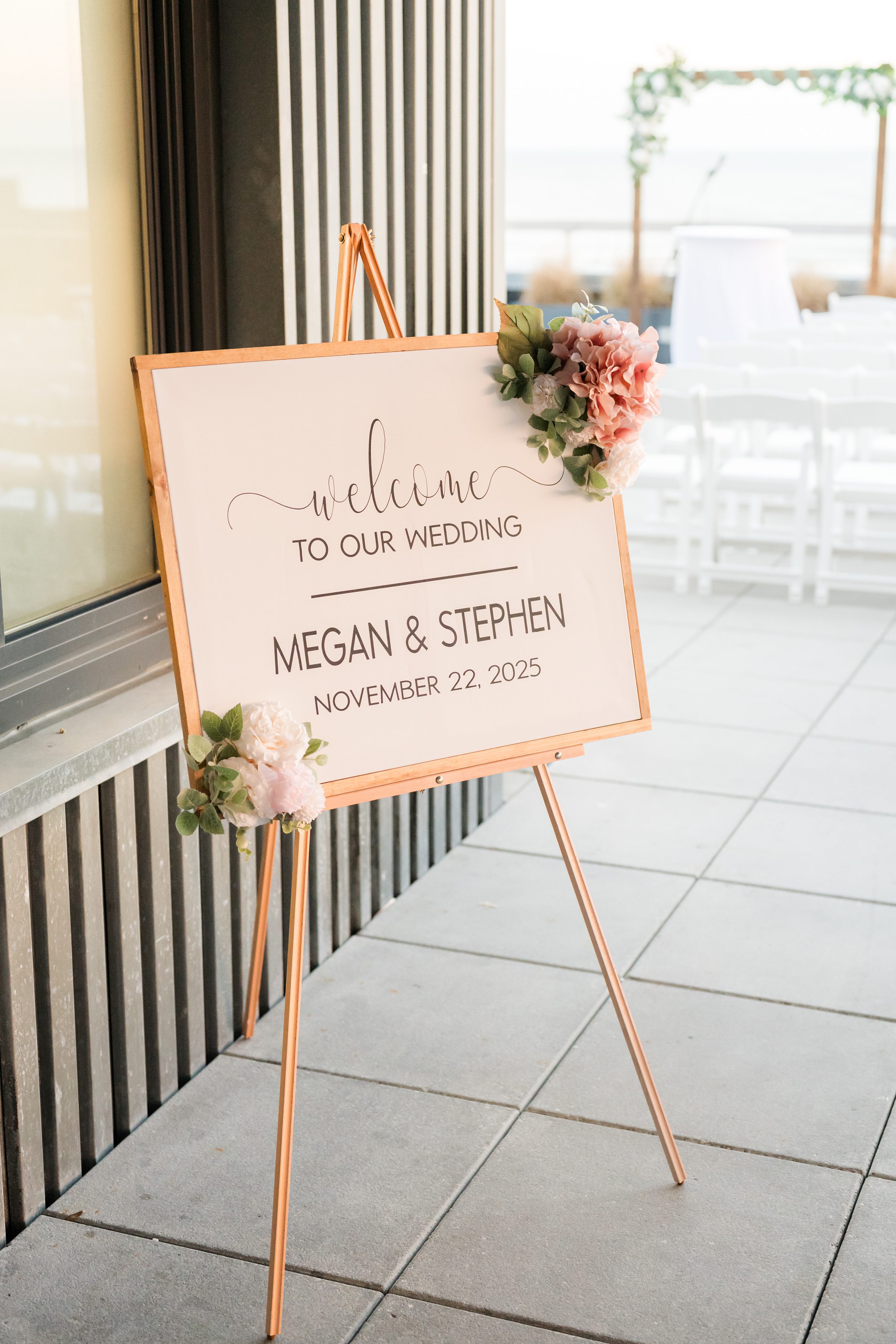 Wedding welcome sign on an easel reading “Megan & Stephen, November 22, 2025,” decorated with soft floral accents at a rooftop venue.