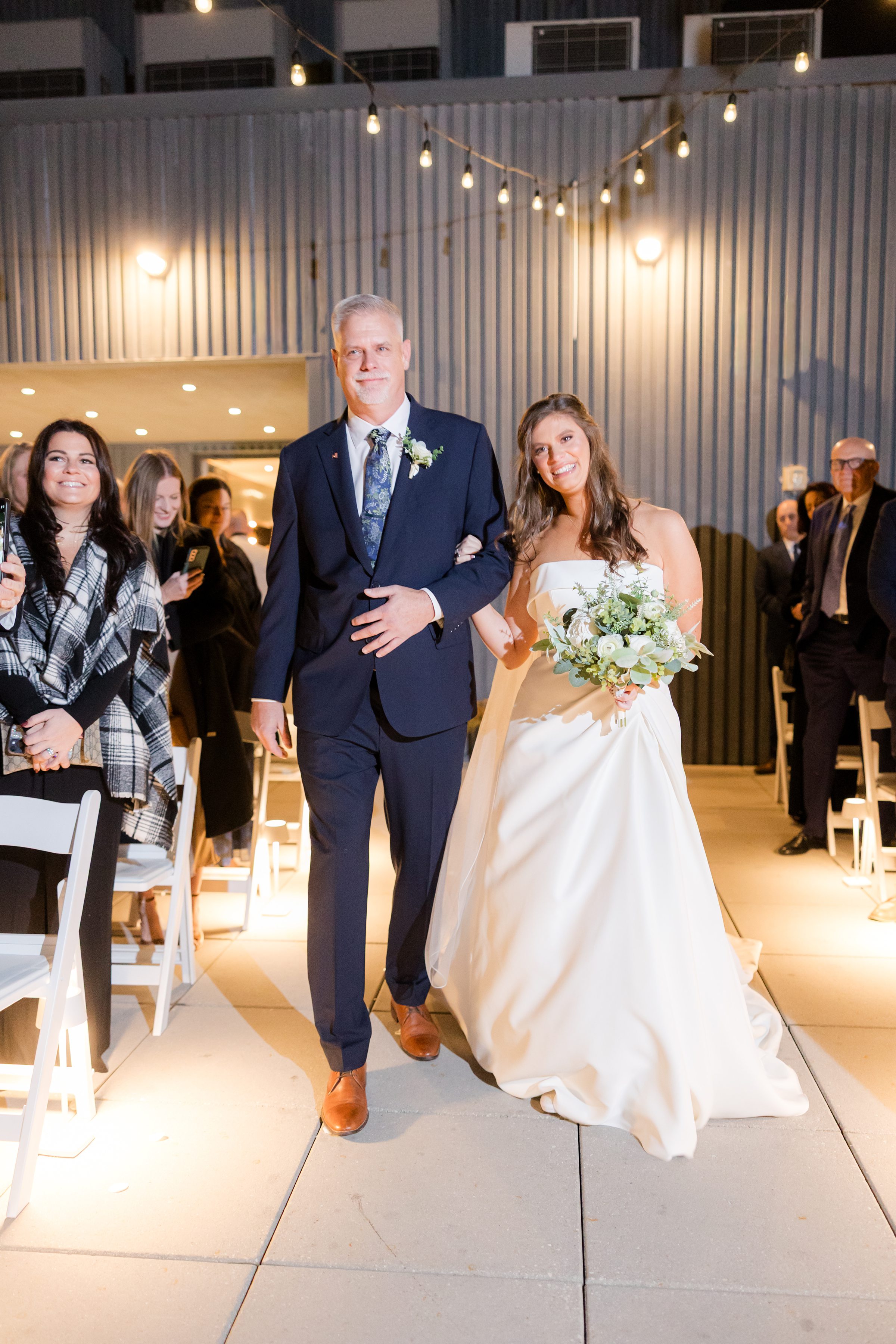 Bride walks down the aisle with her father, smiling and holding her bouquet as guests look on under warm string lights.