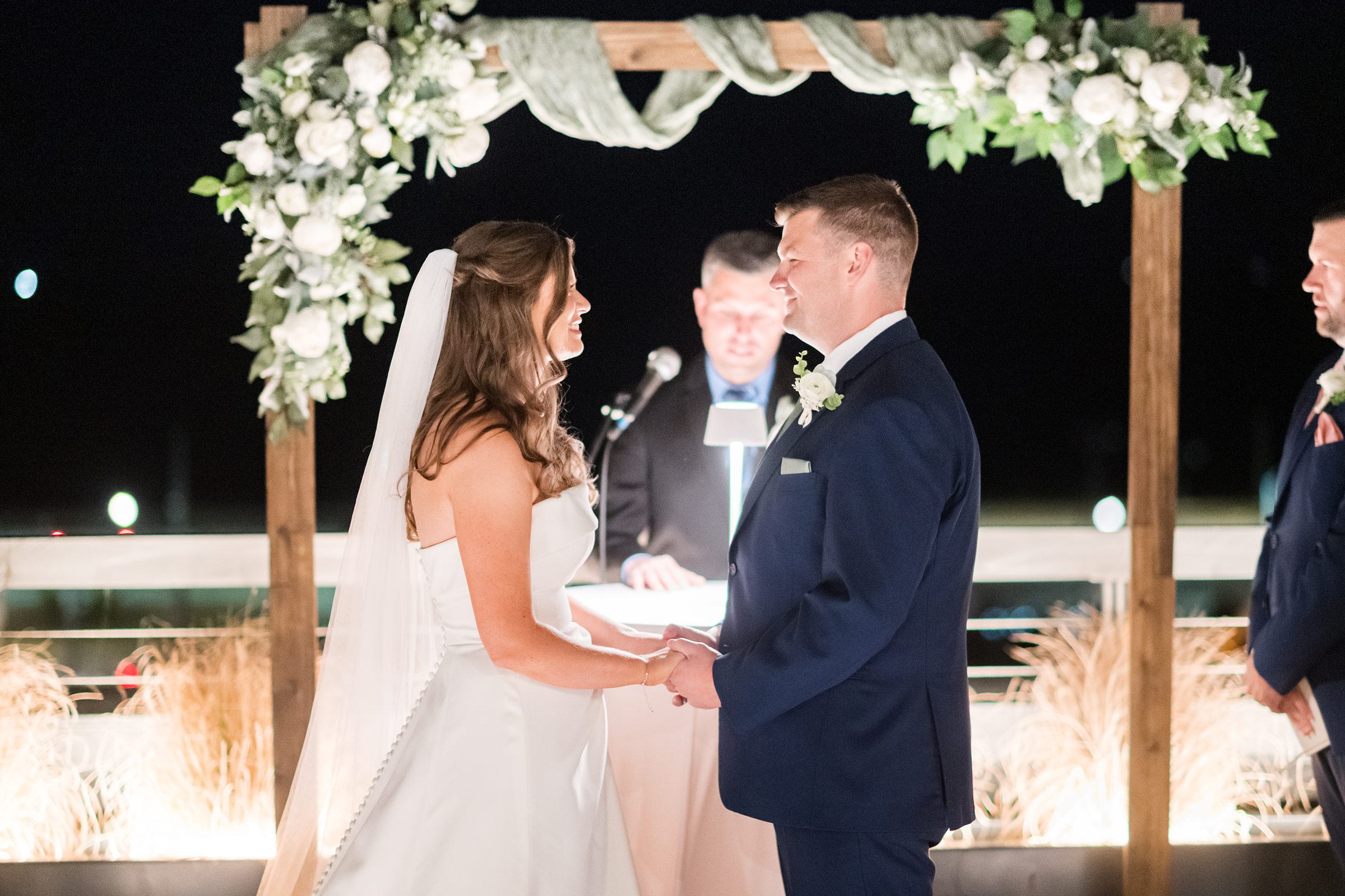 Bride and groom smiling at each other while holding hands during the ceremony, officiant standing behind them.