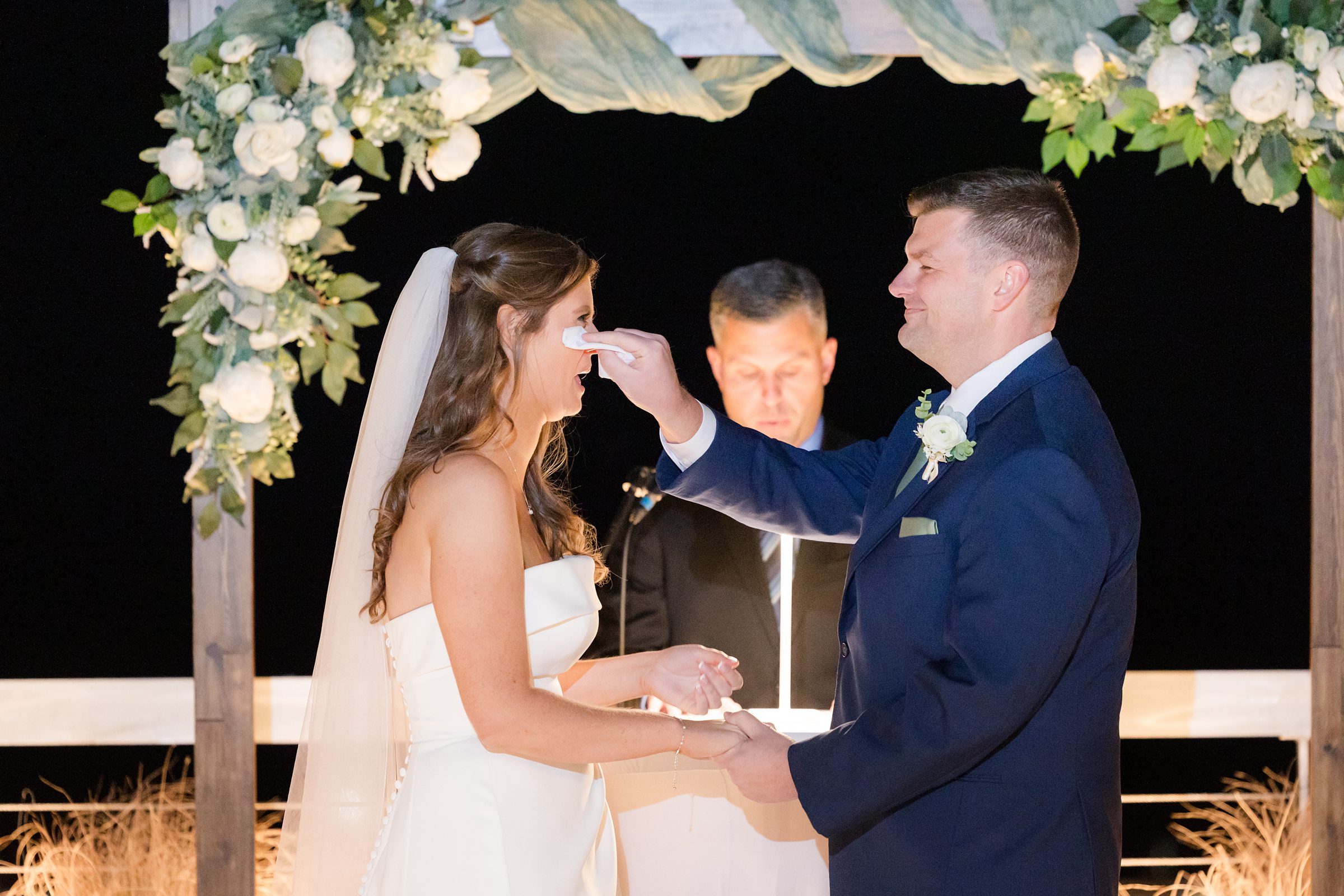 Groom gently wiping tears from the bride’s face during their vows under a flower-decorated arch at night.