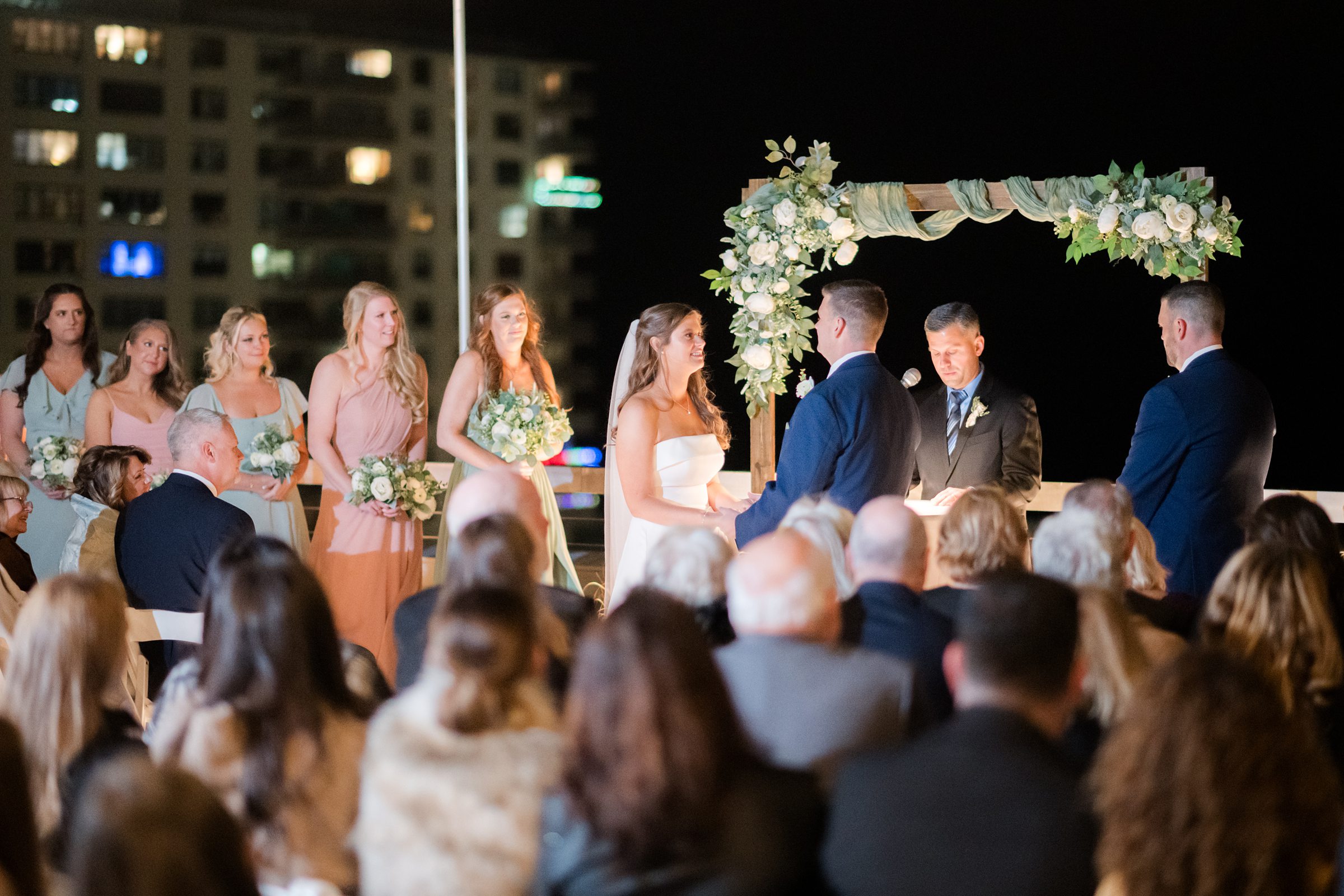 Bride and groom stand together beneath a softly lit floral arch, surrounded by loved ones, sharing a heartfelt and romantic moment during their evening ceremony.
