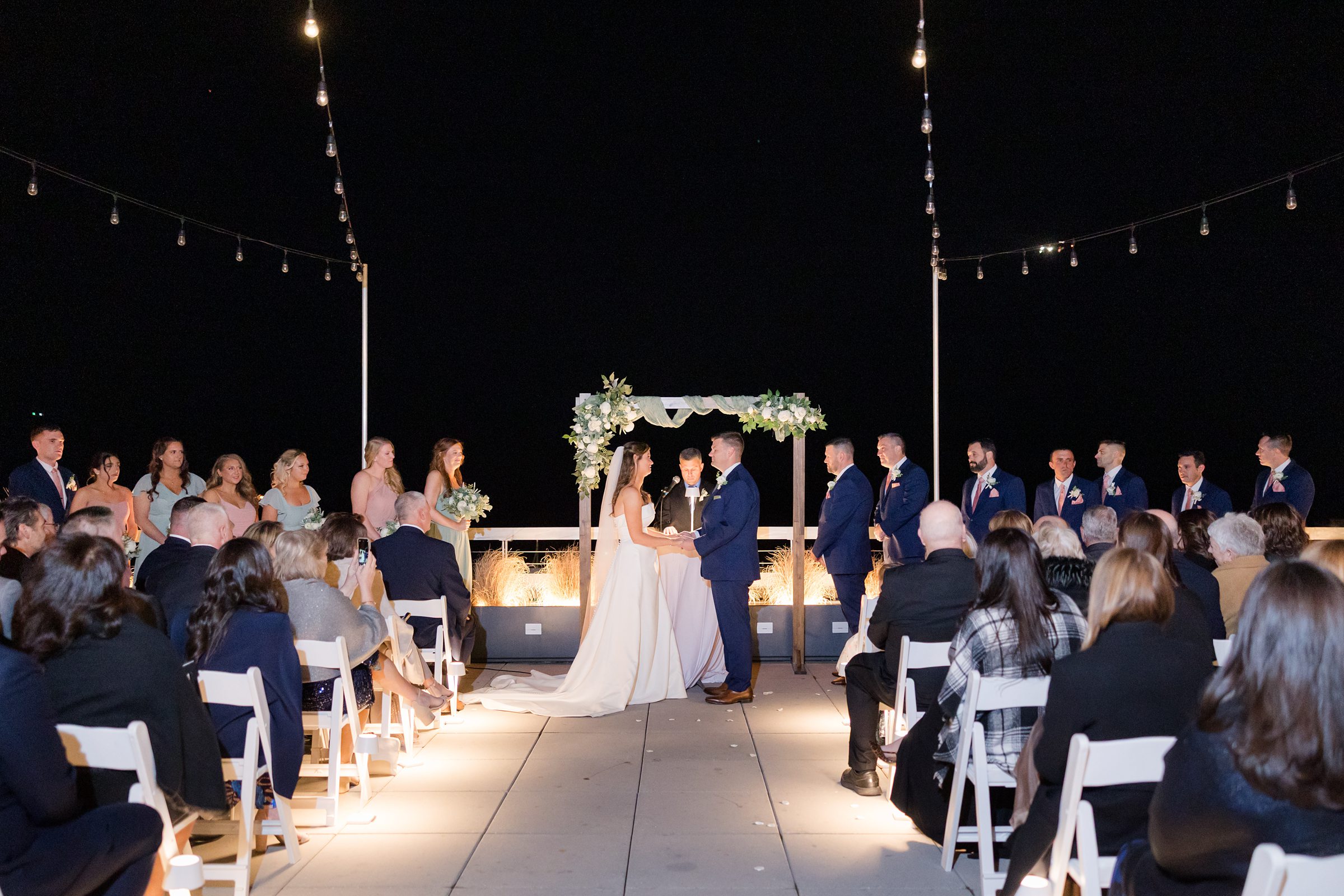 Bride and groom exchange vows beneath a glowing floral arch, surrounded by loved ones, as soft lights and the night sky create a deeply romantic atmosphere.