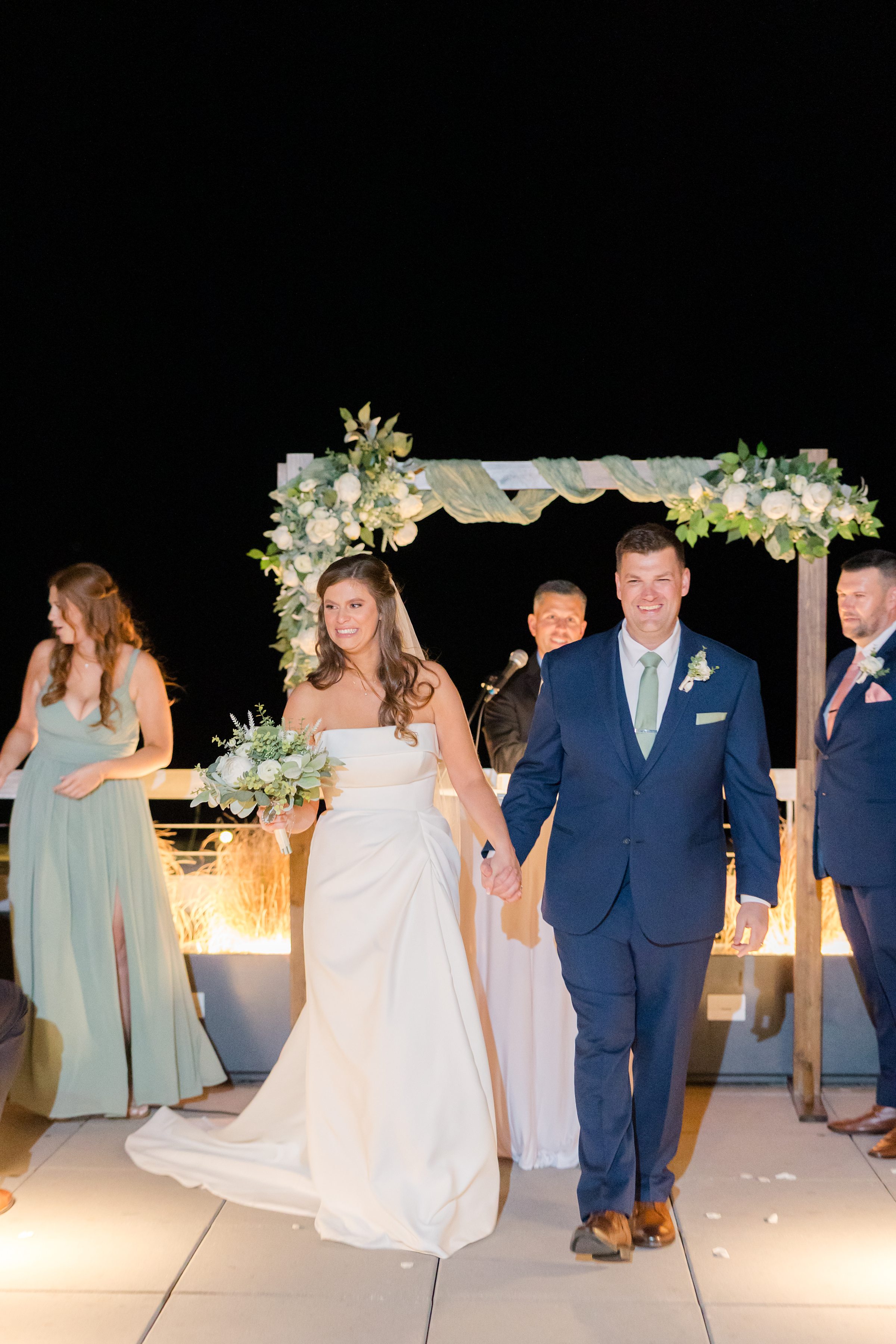 Bride and groom smiling and walking together under a floral arch at an evening wedding ceremony.
