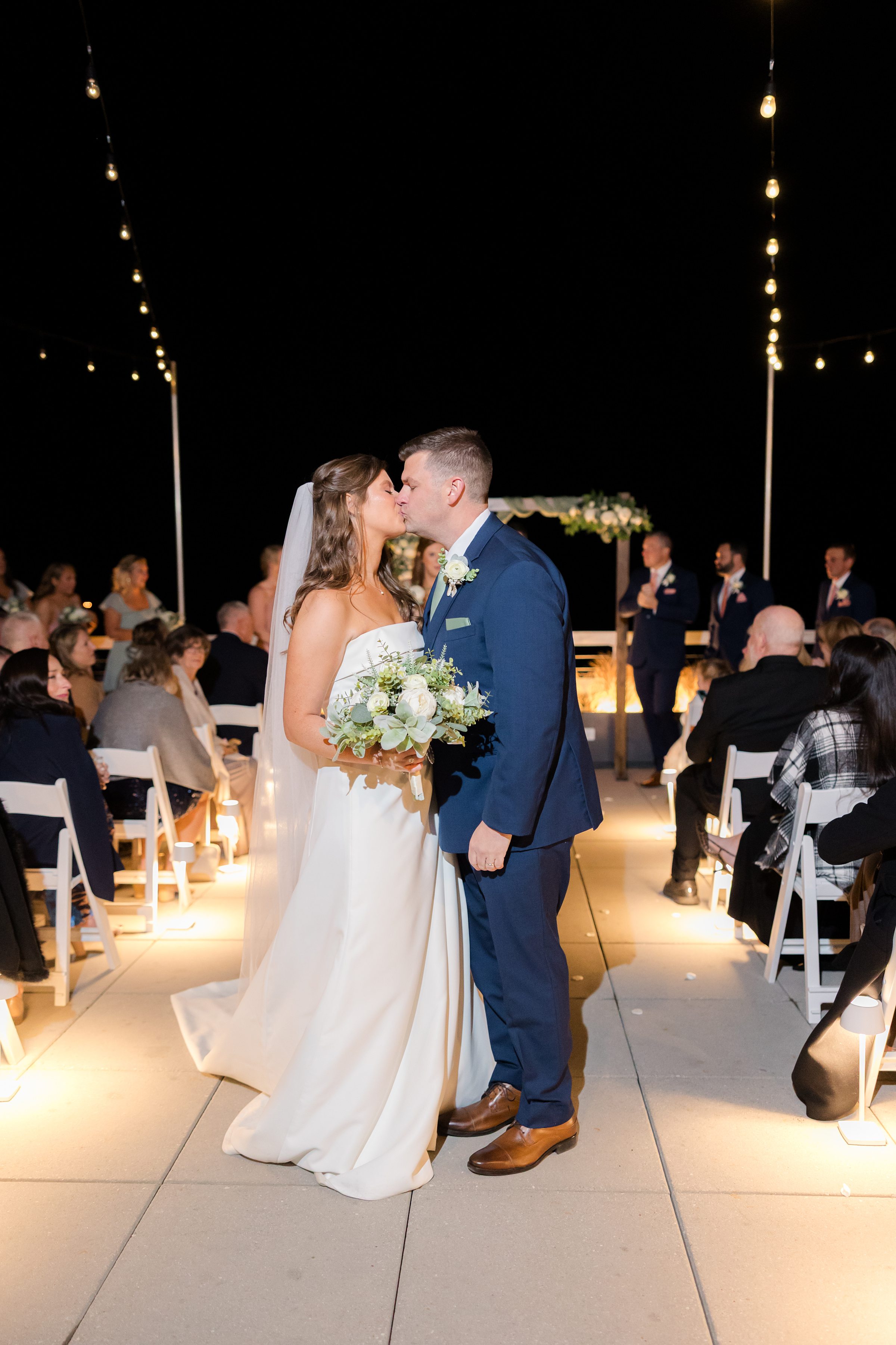 Bride and groom share a romantic kiss beneath glowing string lights, surrounded by loved ones at their evening wedding ceremony.