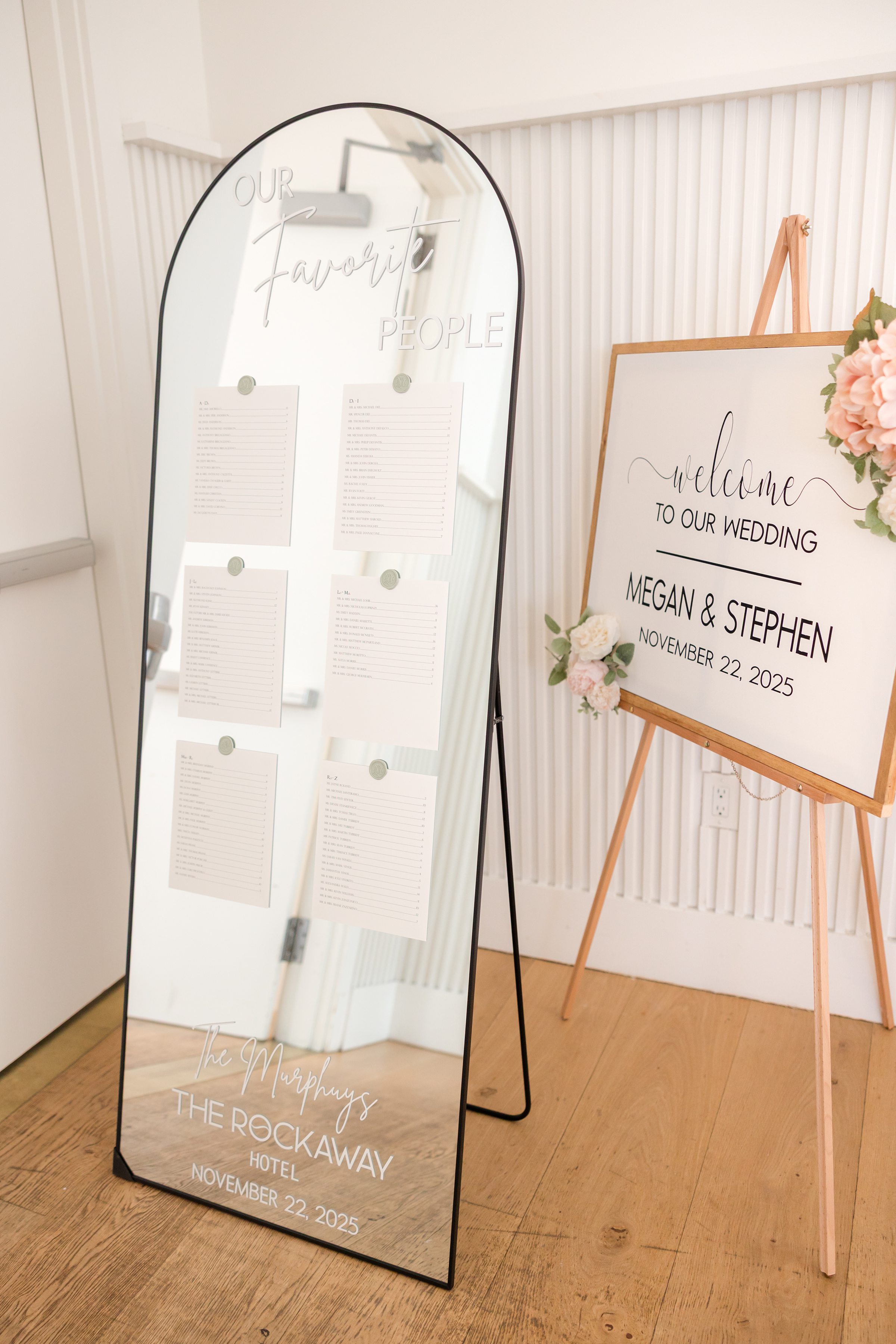 Wedding welcome sign and seating chart displayed on a mirror and easel with floral accents, featuring the couple’s names and date.