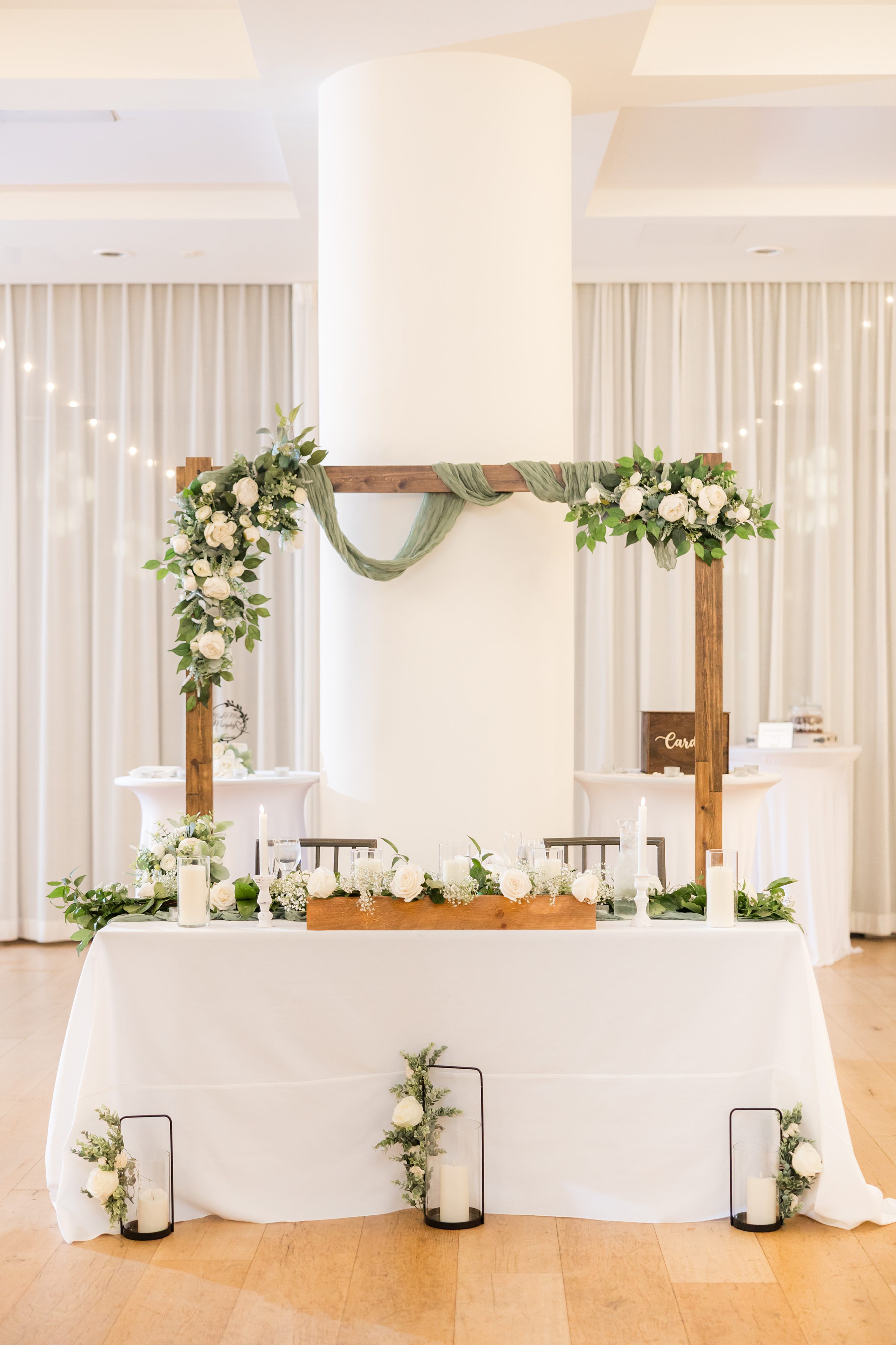 Wooden arch decorated with greenery and white flowers above a table with candles and floral arrangements in a softly lit indoor setting.