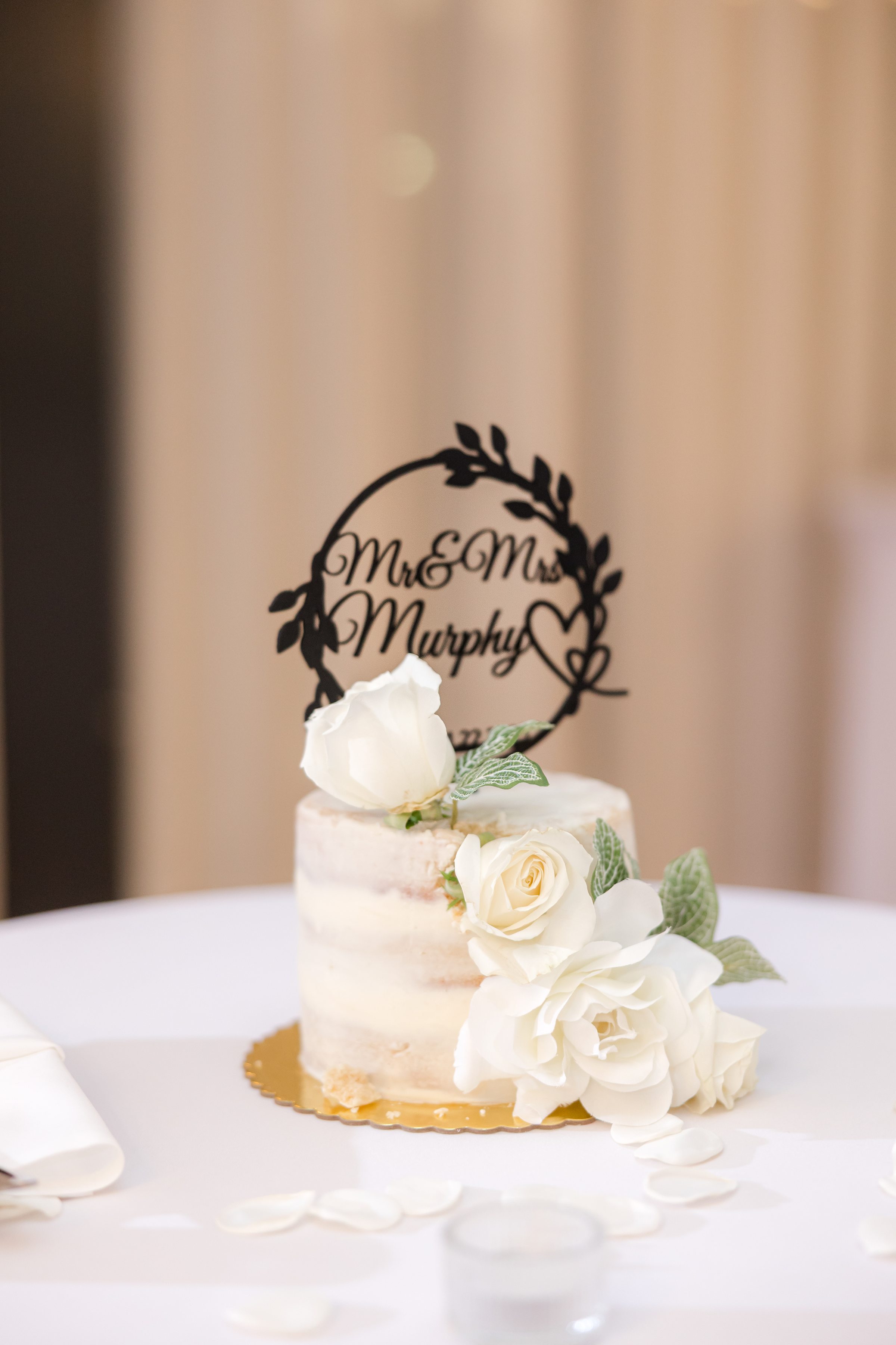 White wedding cake topped with a personalized “Mr & Mrs Murphy” topper and decorated with white roses on a table.