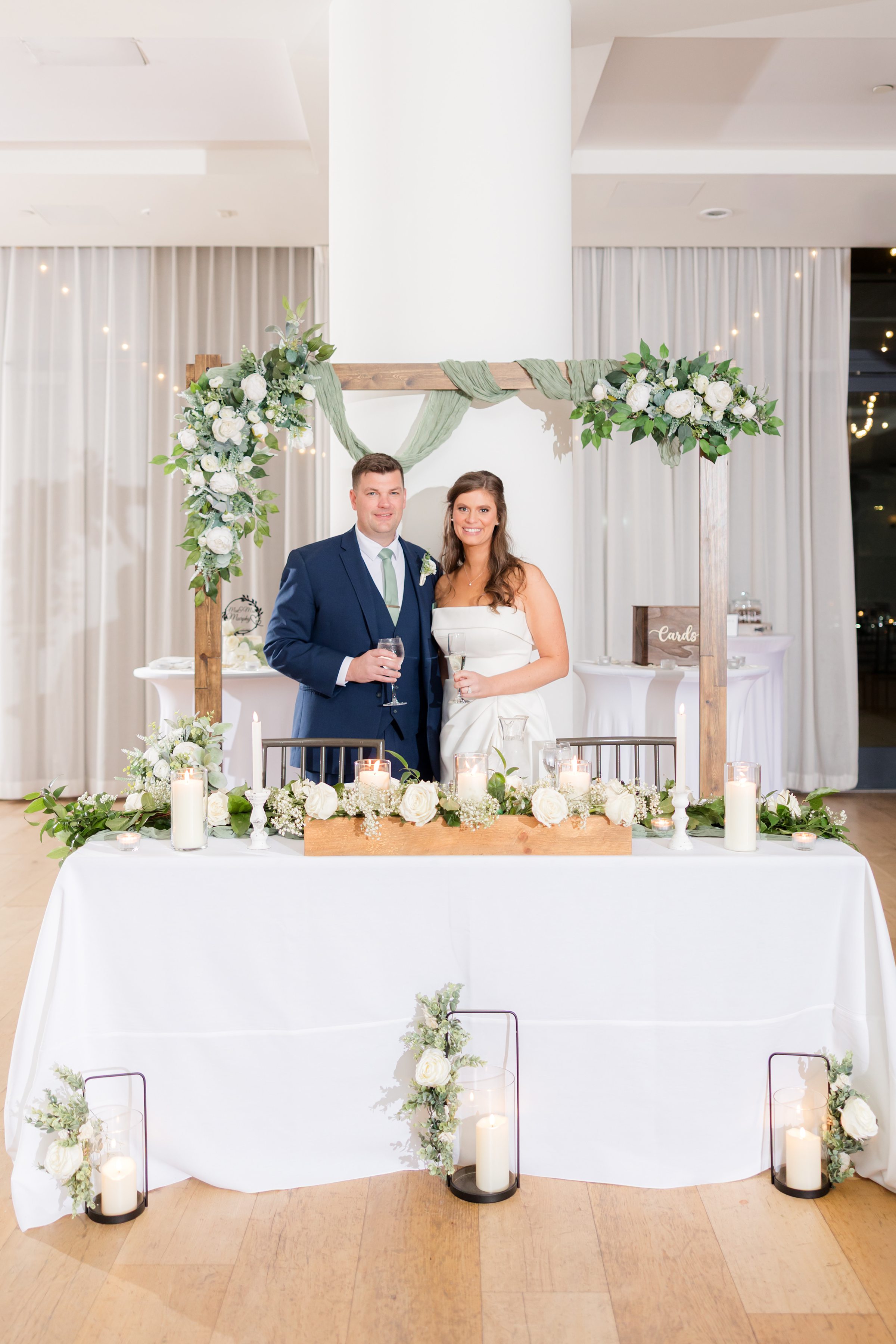 Bride and groom stand behind decorated sweetheart table with candles and greenery, holding drinks and smiling