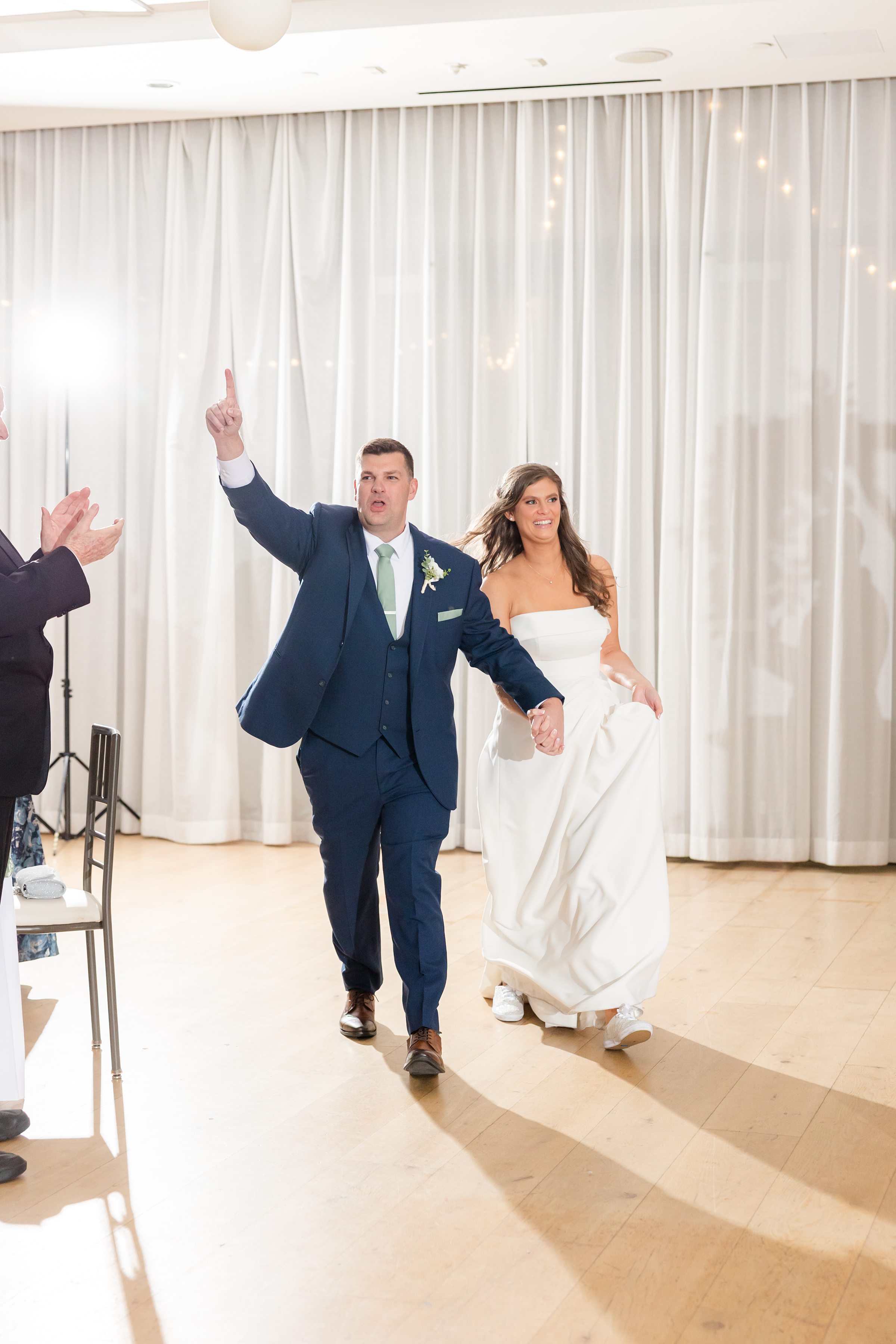 Bride and groom make a joyful entrance, holding hands as he raises his arm in celebration during their wedding reception.