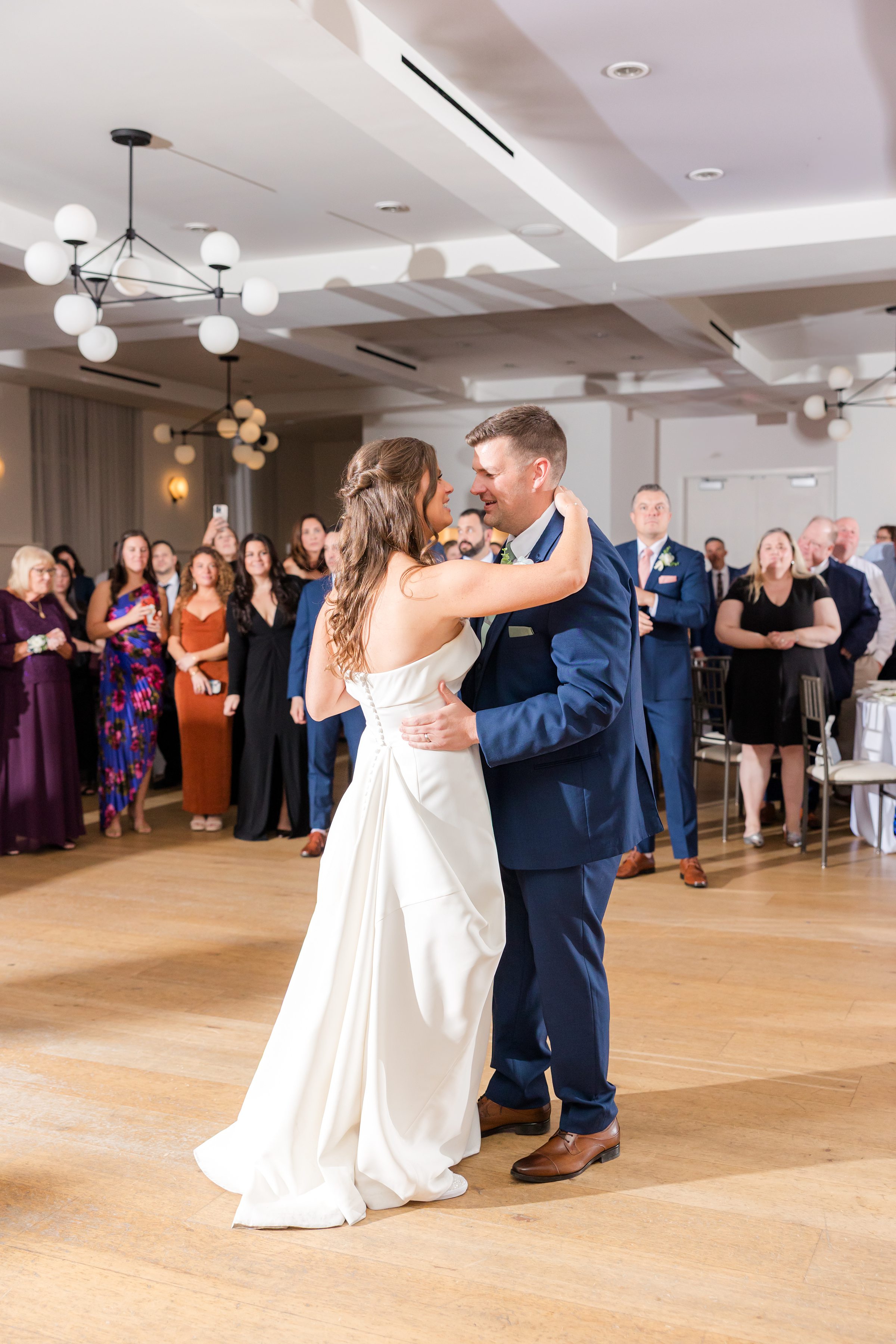 Bride and groom slow dance in the center of a large room while guests stand in a circle watching.