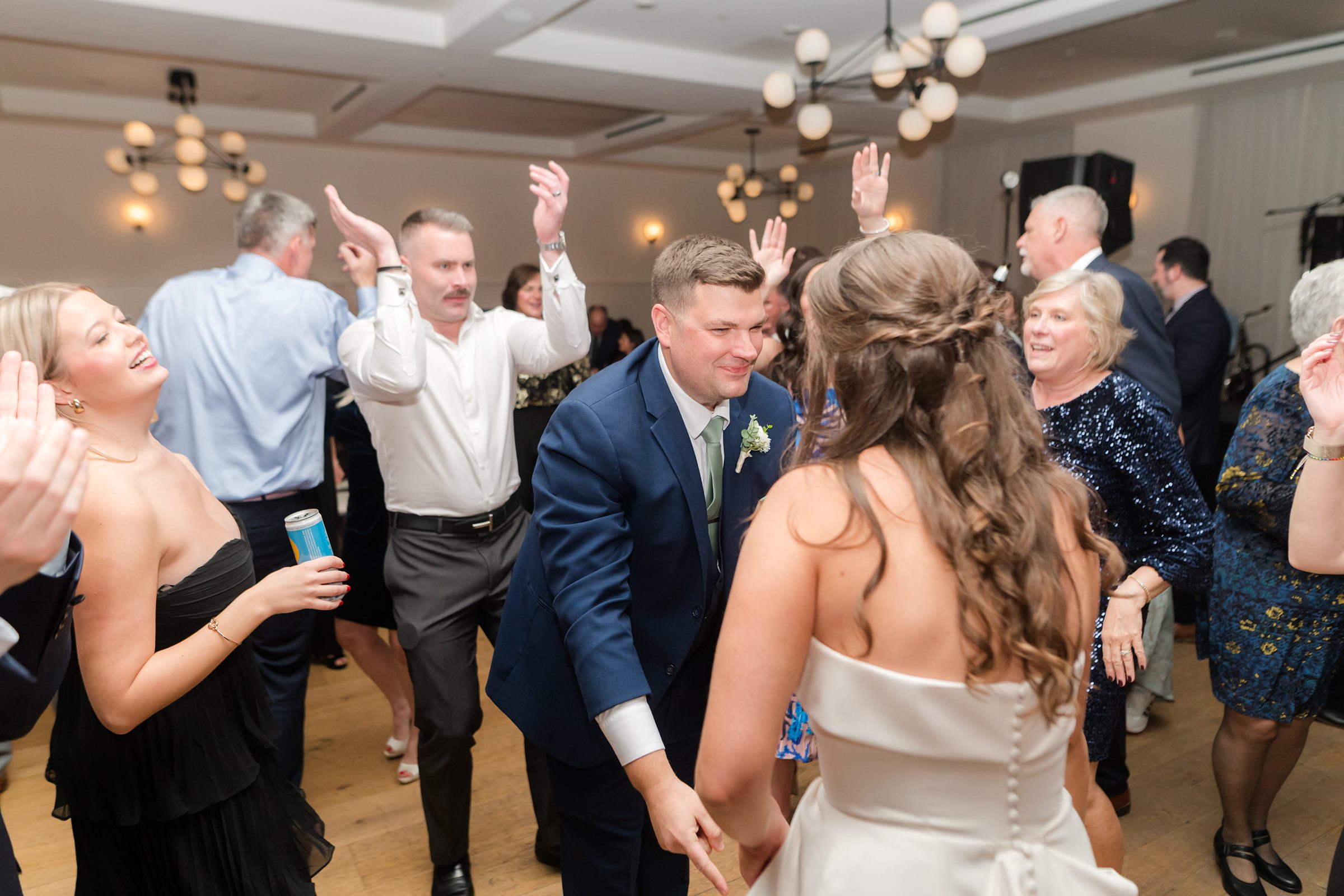 Bride and groom dance together among guests, with people clapping and raising hands around them.