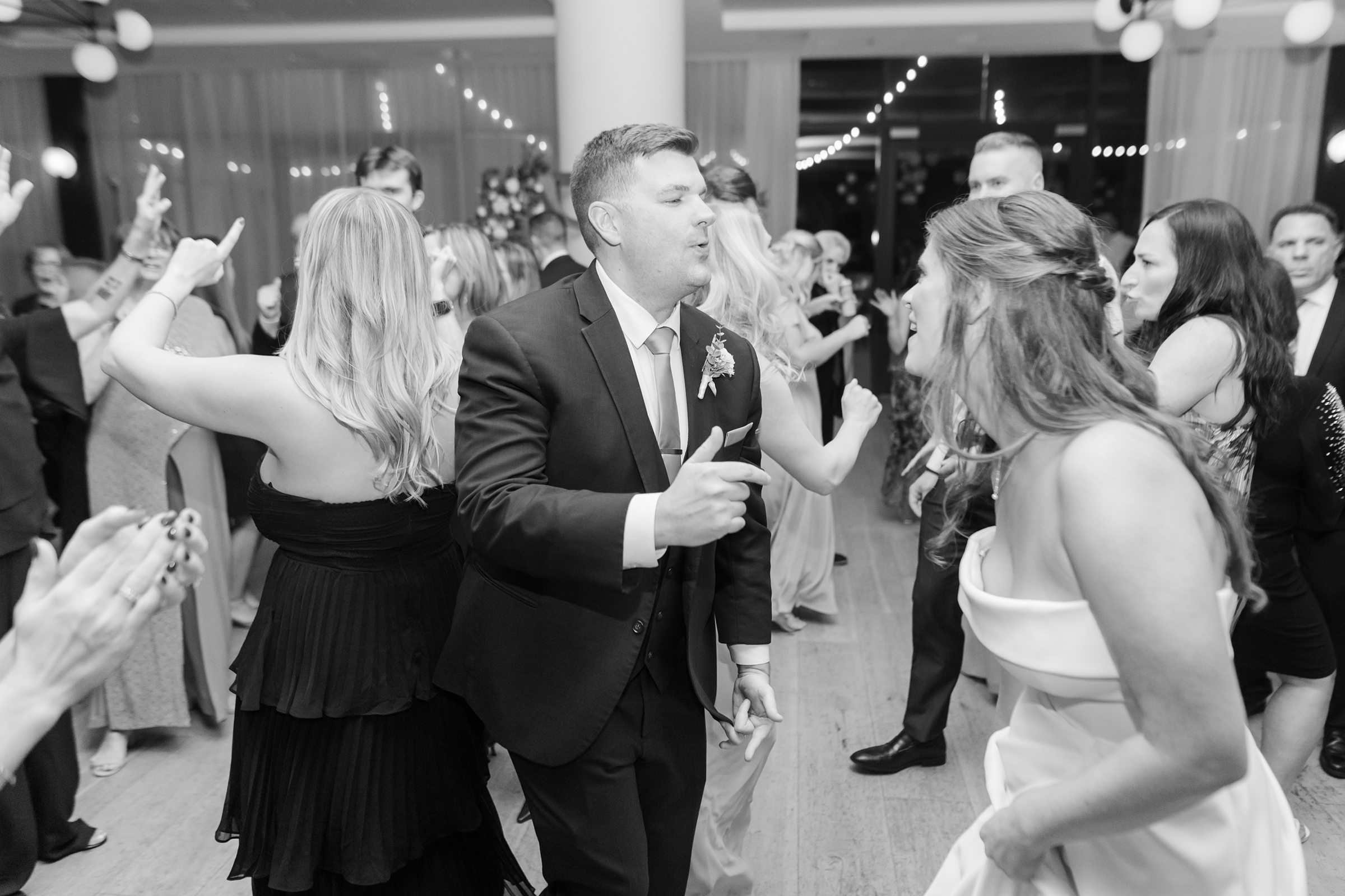 Bride and groom laugh and dance together surrounded by guests during a lively wedding reception.