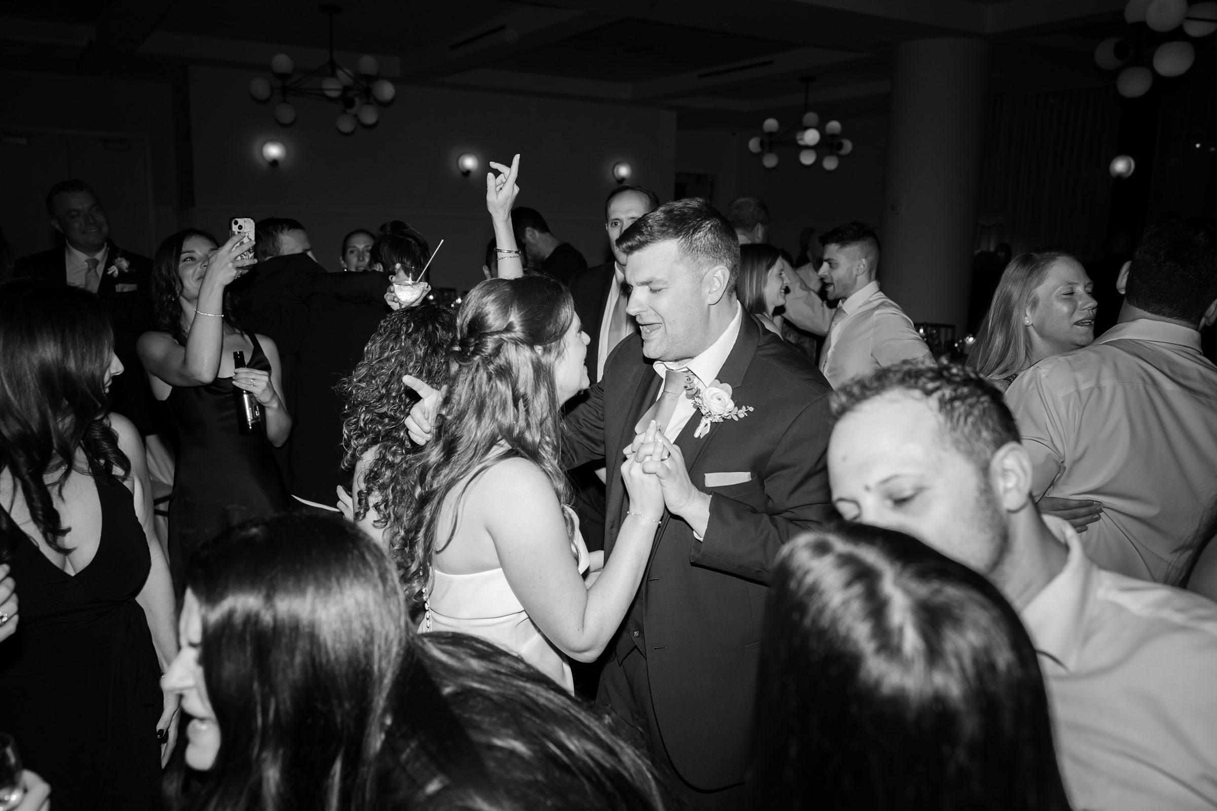 Bride and groom hold hands and sway together on the dance floor, lost in each other while celebrating among loved ones.