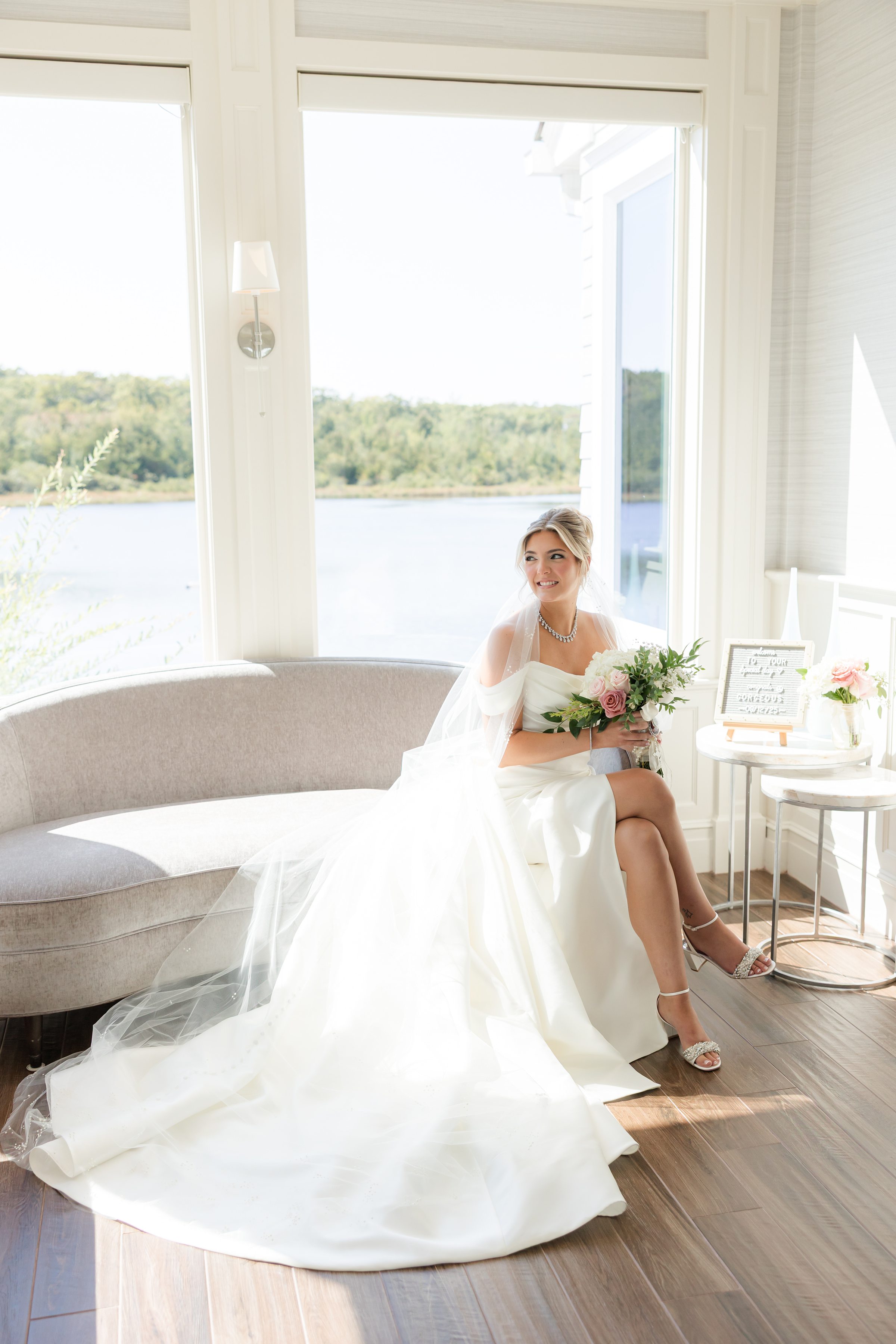 Bride sits by sunlit windows holding her bouquet, gazing out peacefully at the water in a quiet, romantic moment.
