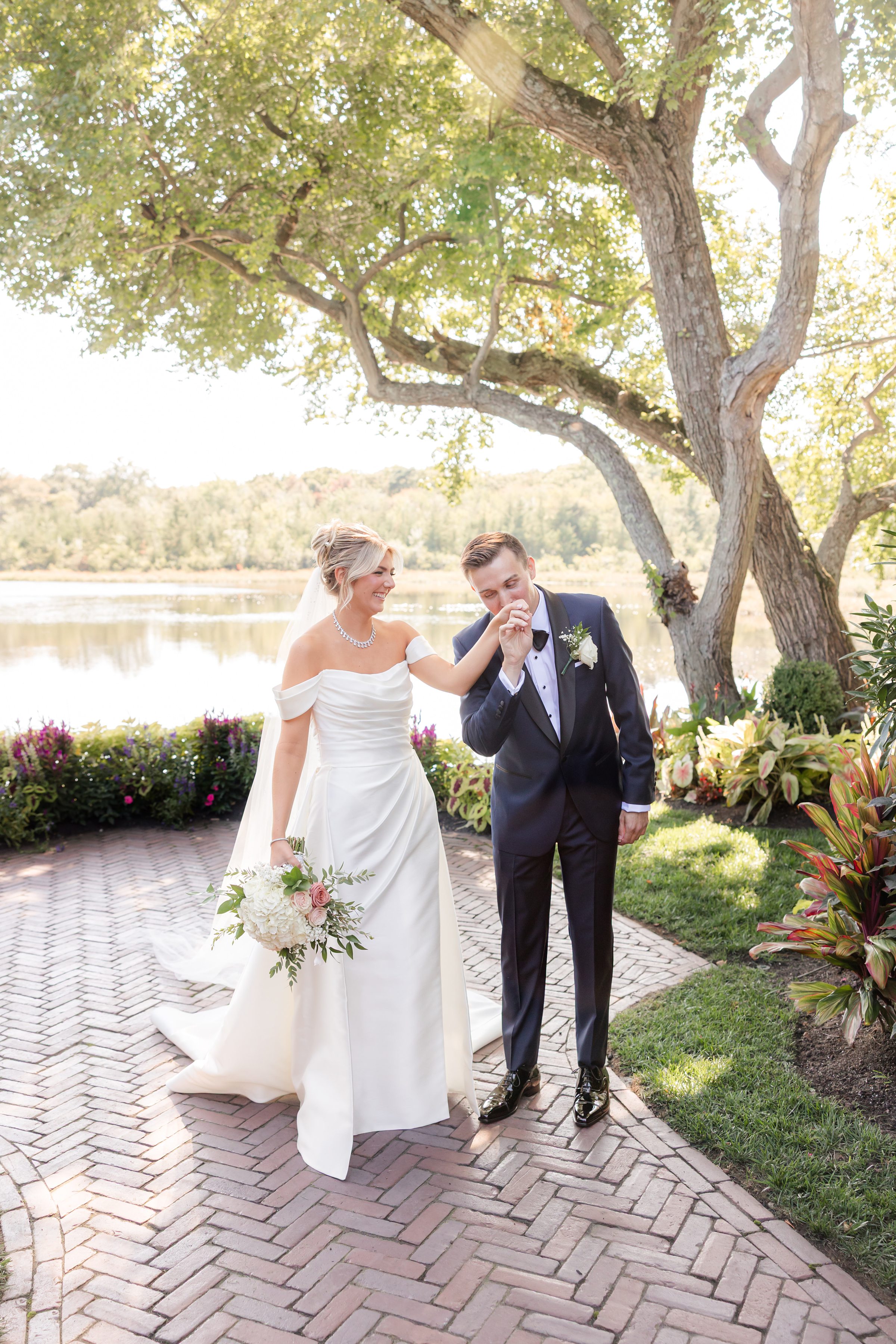 Groom kisses bride’s hand as they walk beside a peaceful lakeside.