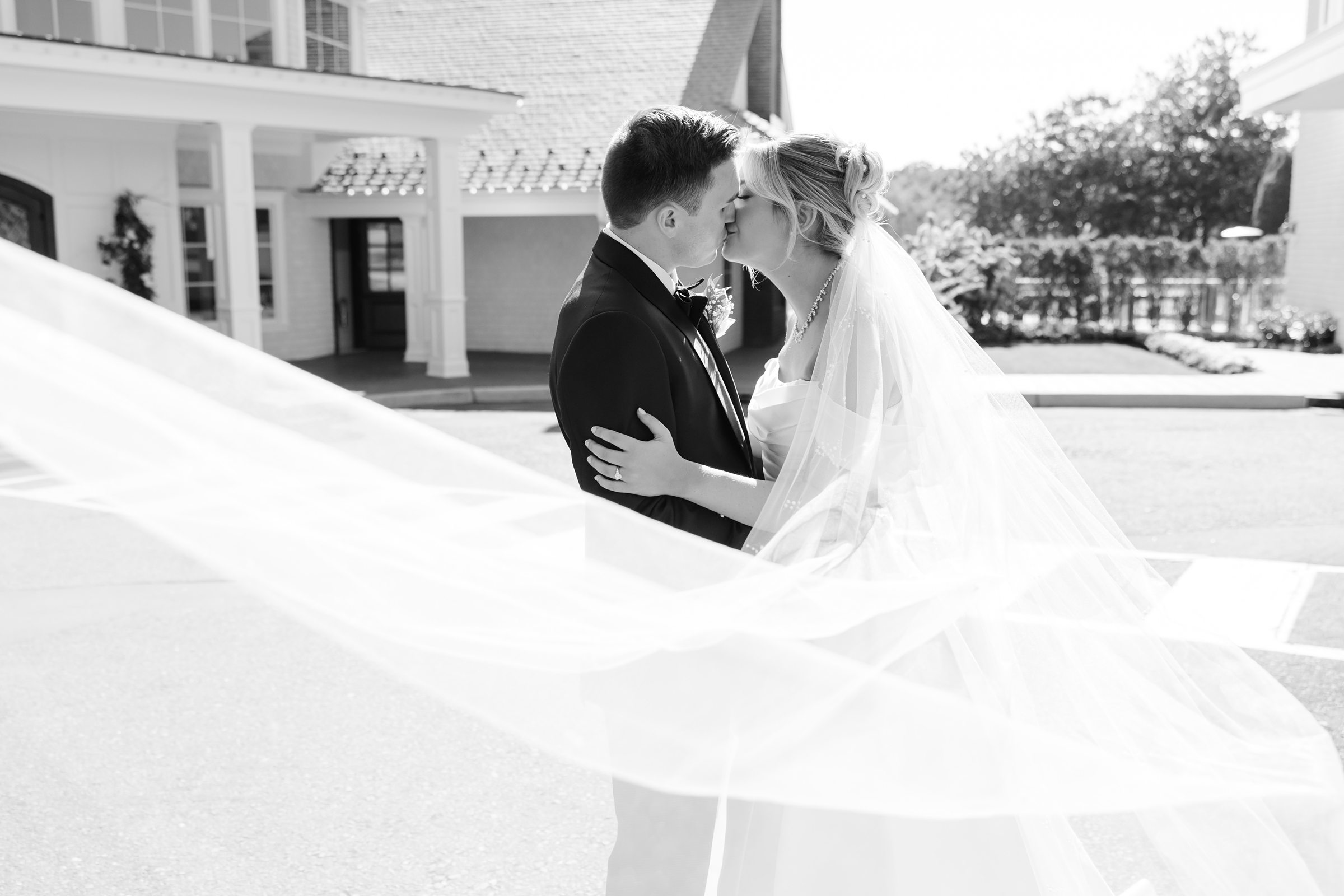 Bride and groom share a soft kiss as her veil flows around them in an elegant black-and-white portrait