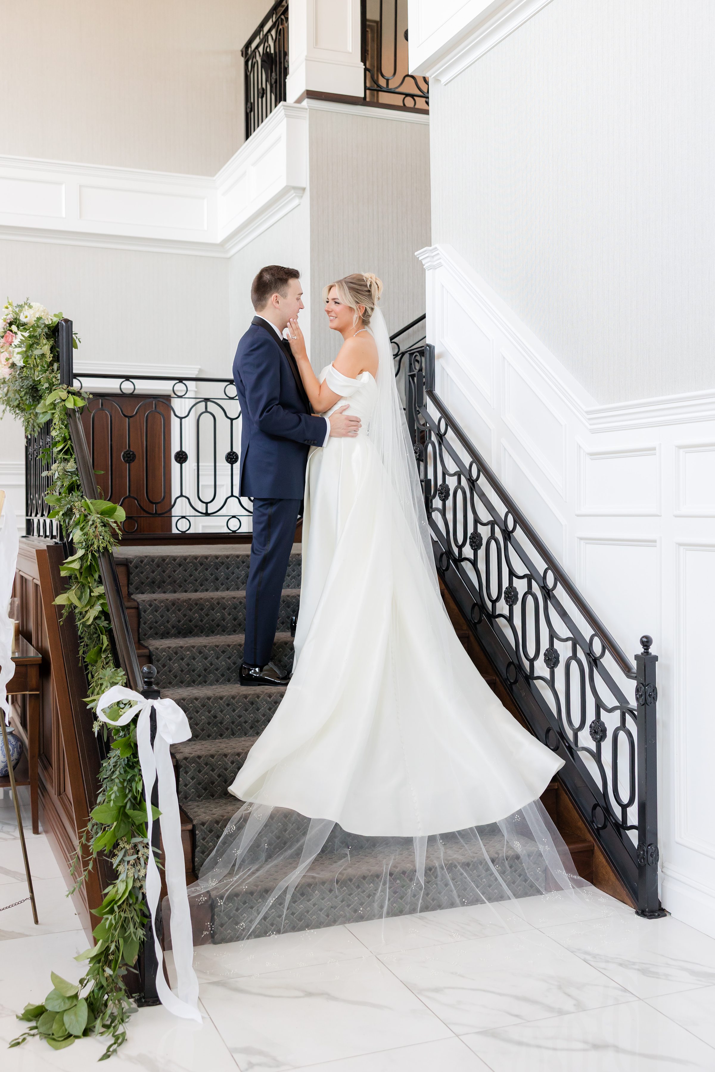 Bride and groom stand close together on a staircase, sharing a soft, intimate look in a bright interior space.