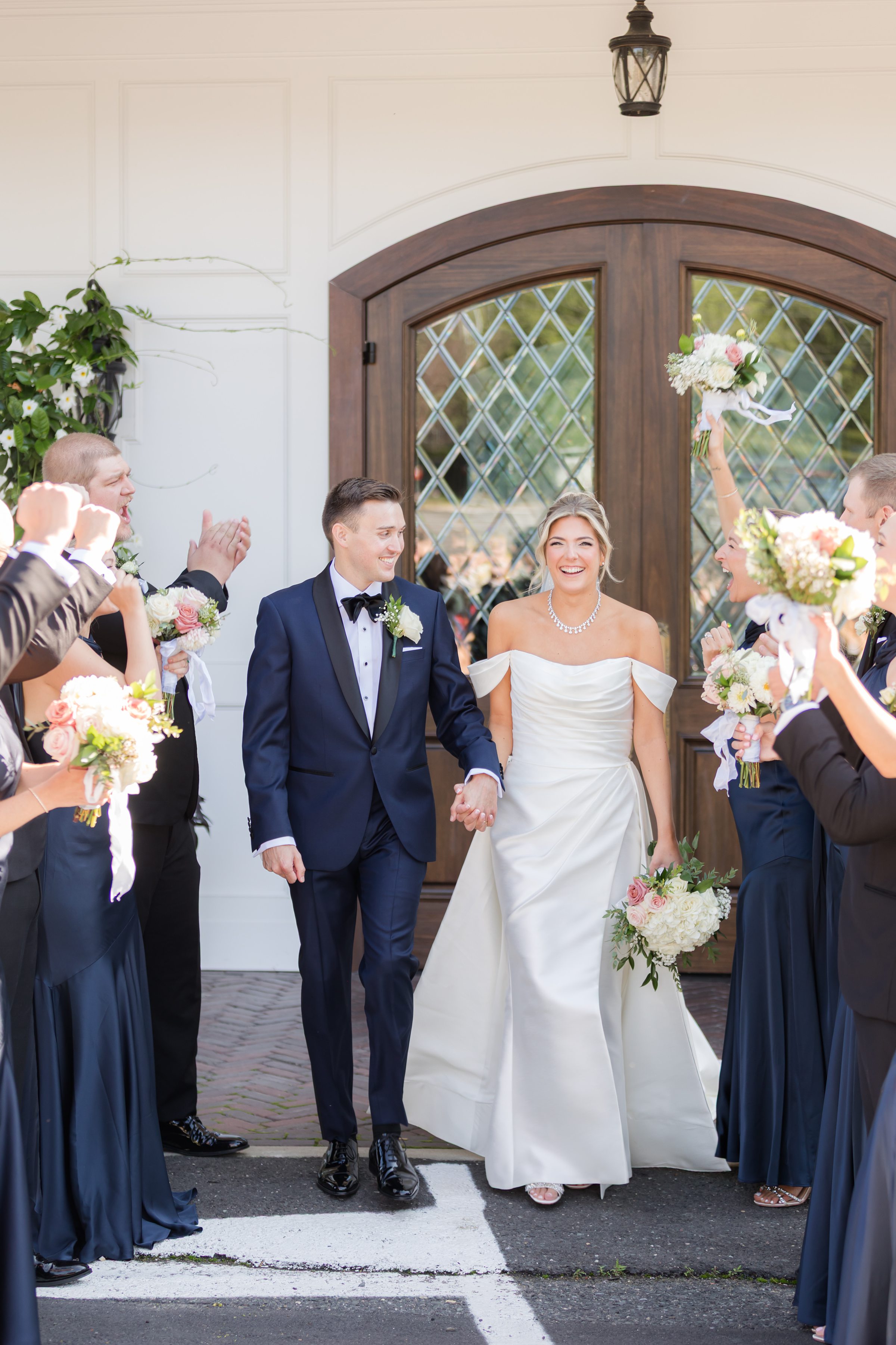 Newlyweds celebrate with their wedding party outside a bright white venue, holding bouquets and cheering.