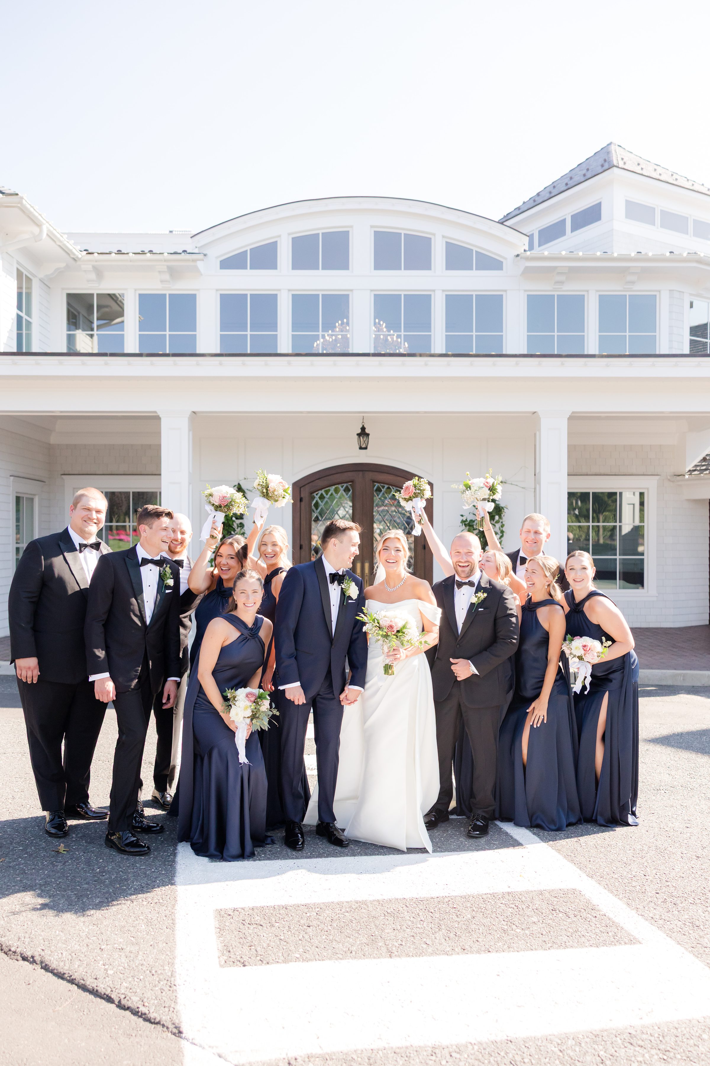 Wedding party celebrates outside a white venue, surrounding the bride and groom as they smile and hold bouquets in bright sunlight.