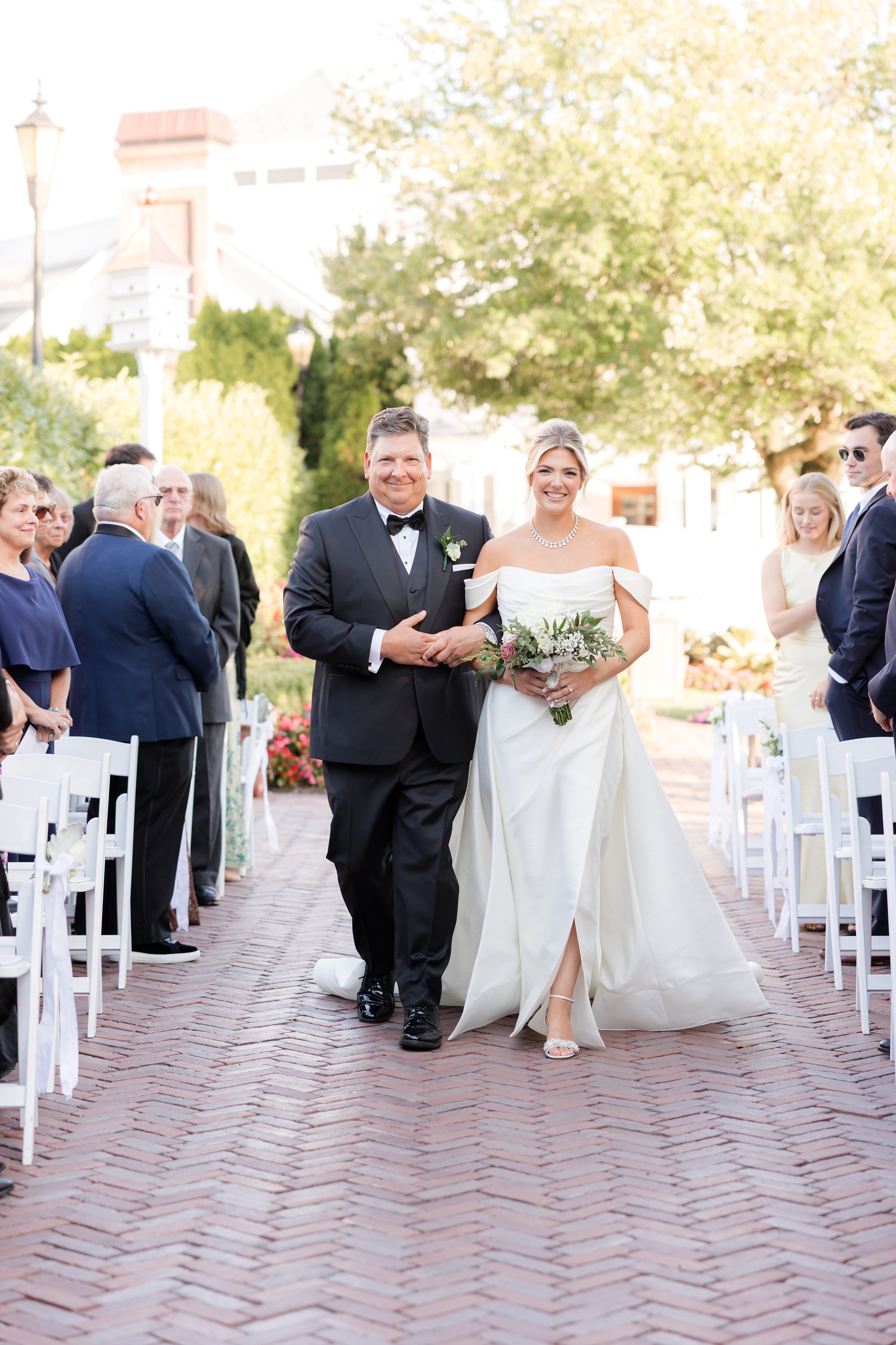 Bride walks down the aisle with her father, smiling brightly as guests look on.