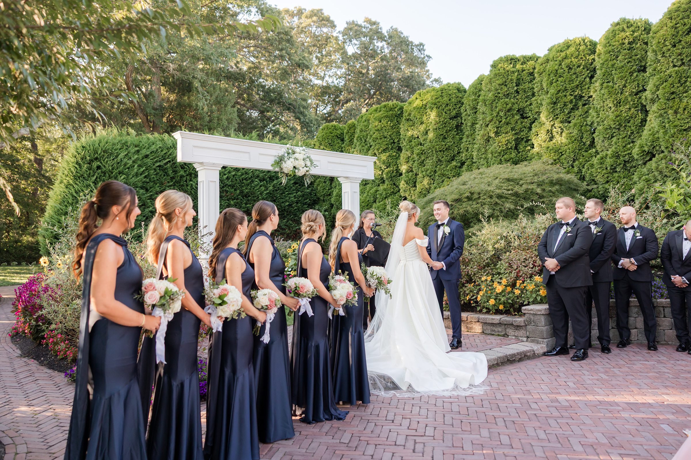 Wedding party stands with the couple during the ceremony, surrounded by greenery and blooming flowers.