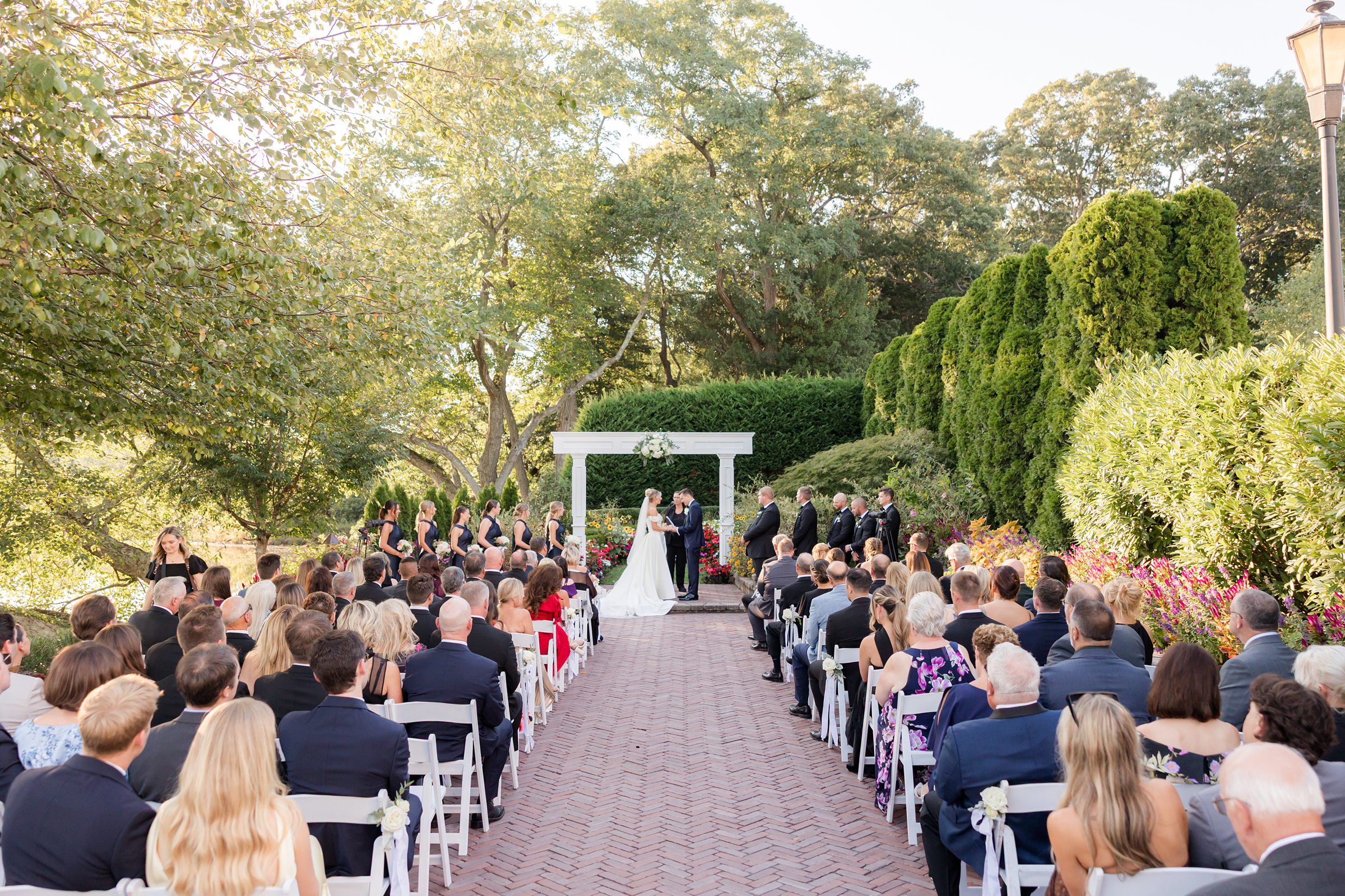 Wide view of an outdoor wedding ceremony with guests seated as the couple exchanges vows beneath a white arbor