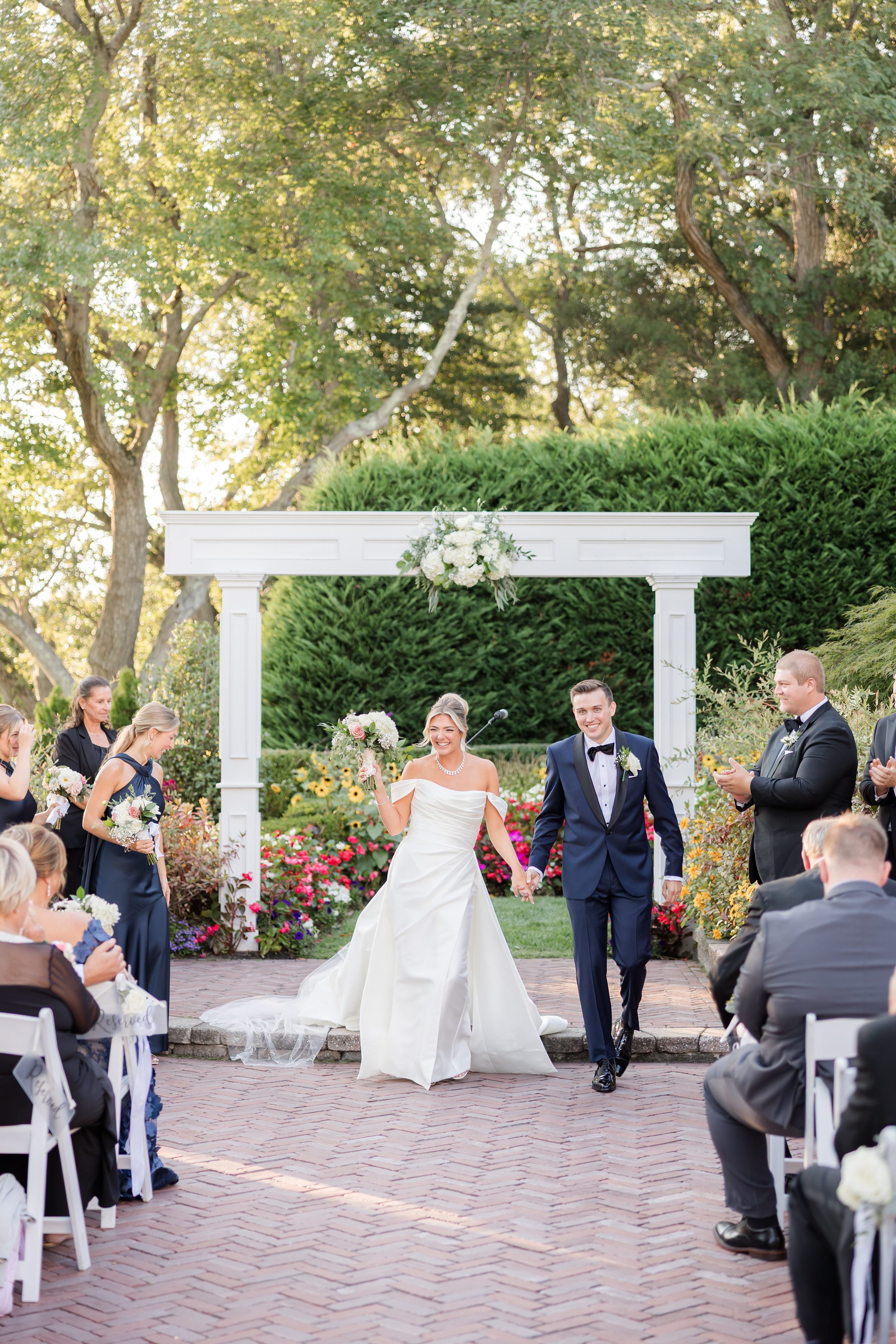 Bride and groom walk hand-in-hand down the aisle, smiling as guests and wedding party applaud beneath a floral garden arbor.