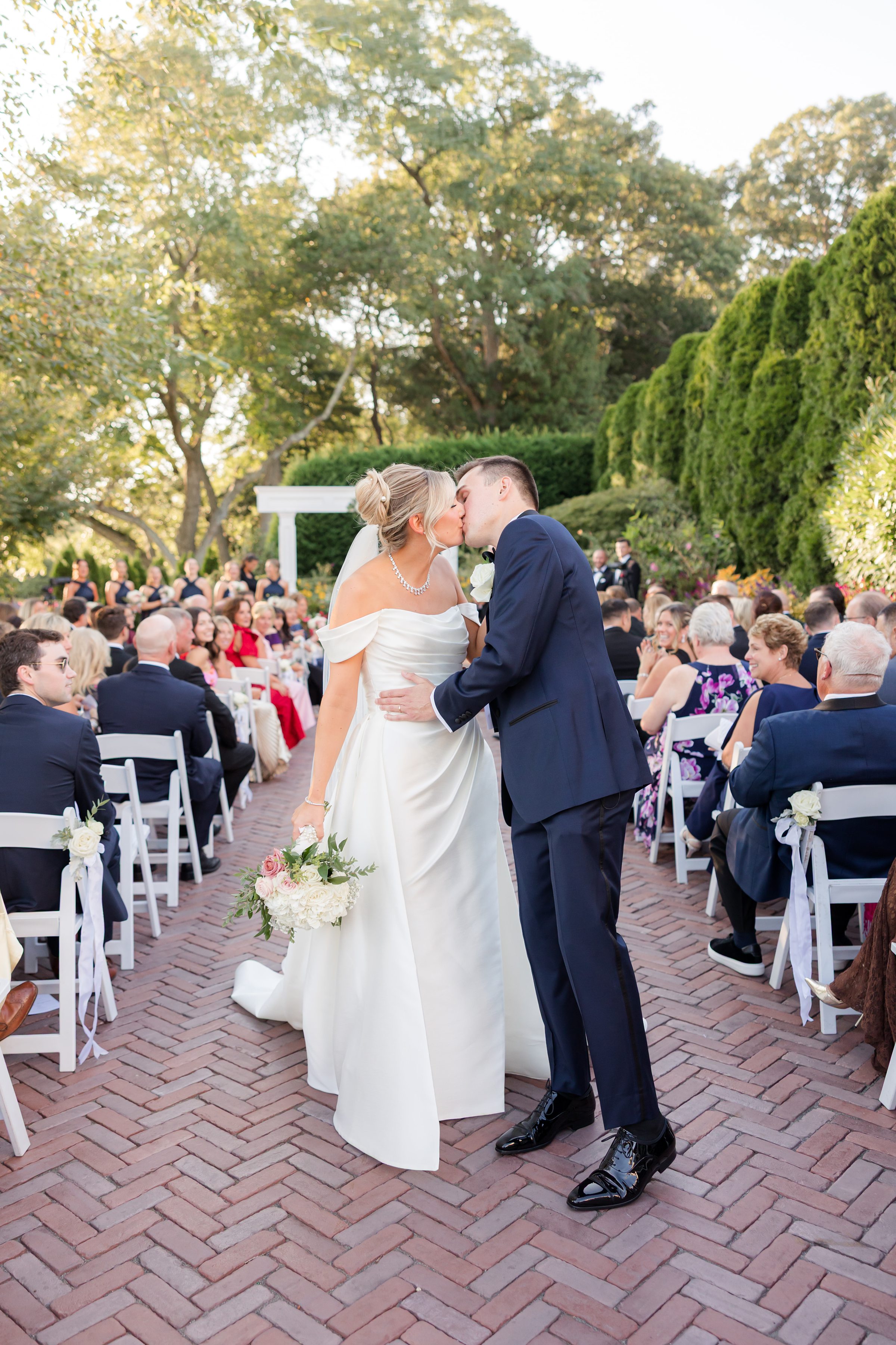 Bride and groom share a kiss in the aisle during their outdoor ceremony, surrounded by seated guests and lush greenery.