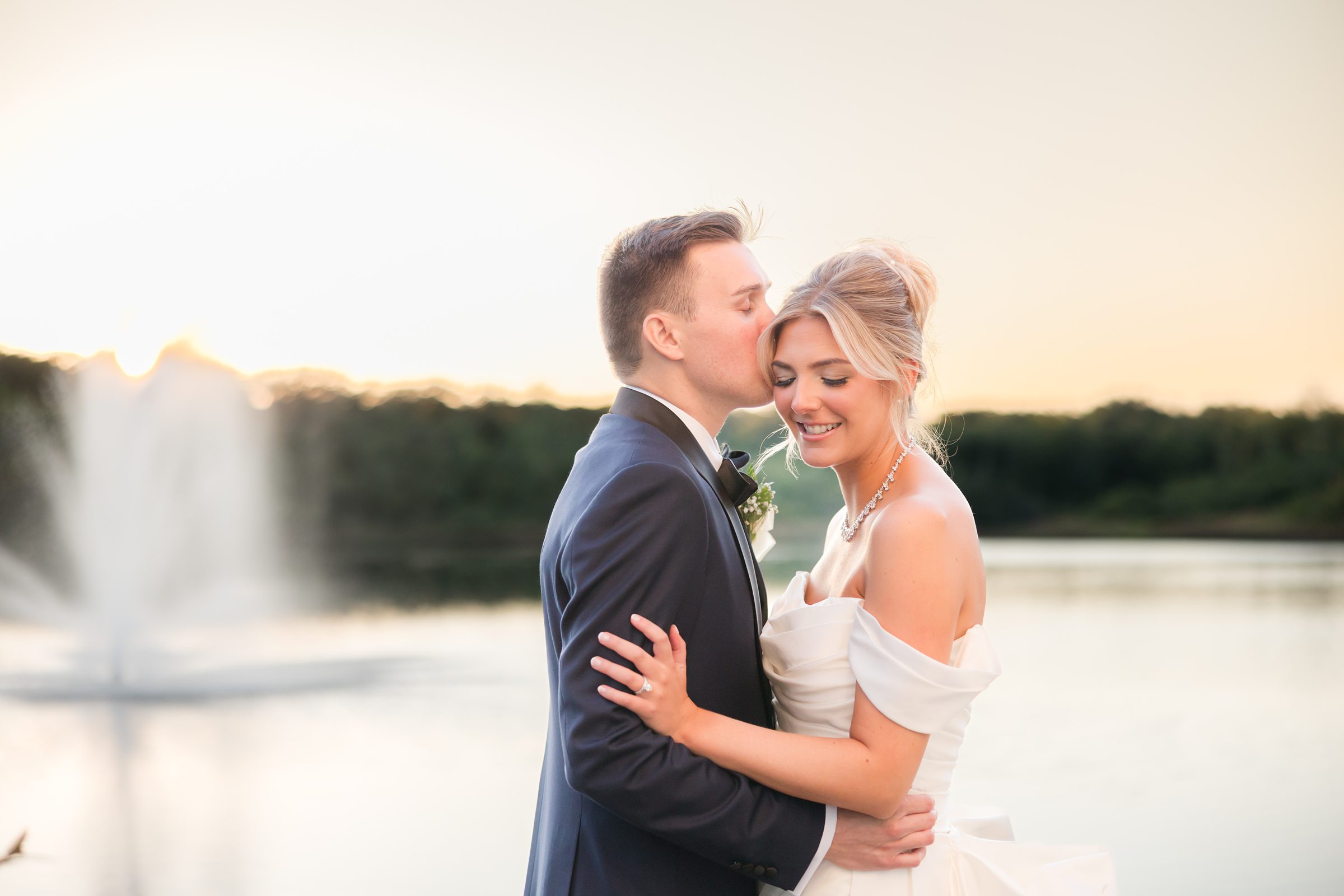 Groom gently kisses bride’s forehead as she smiles, standing by a serene lakeside at sunset.