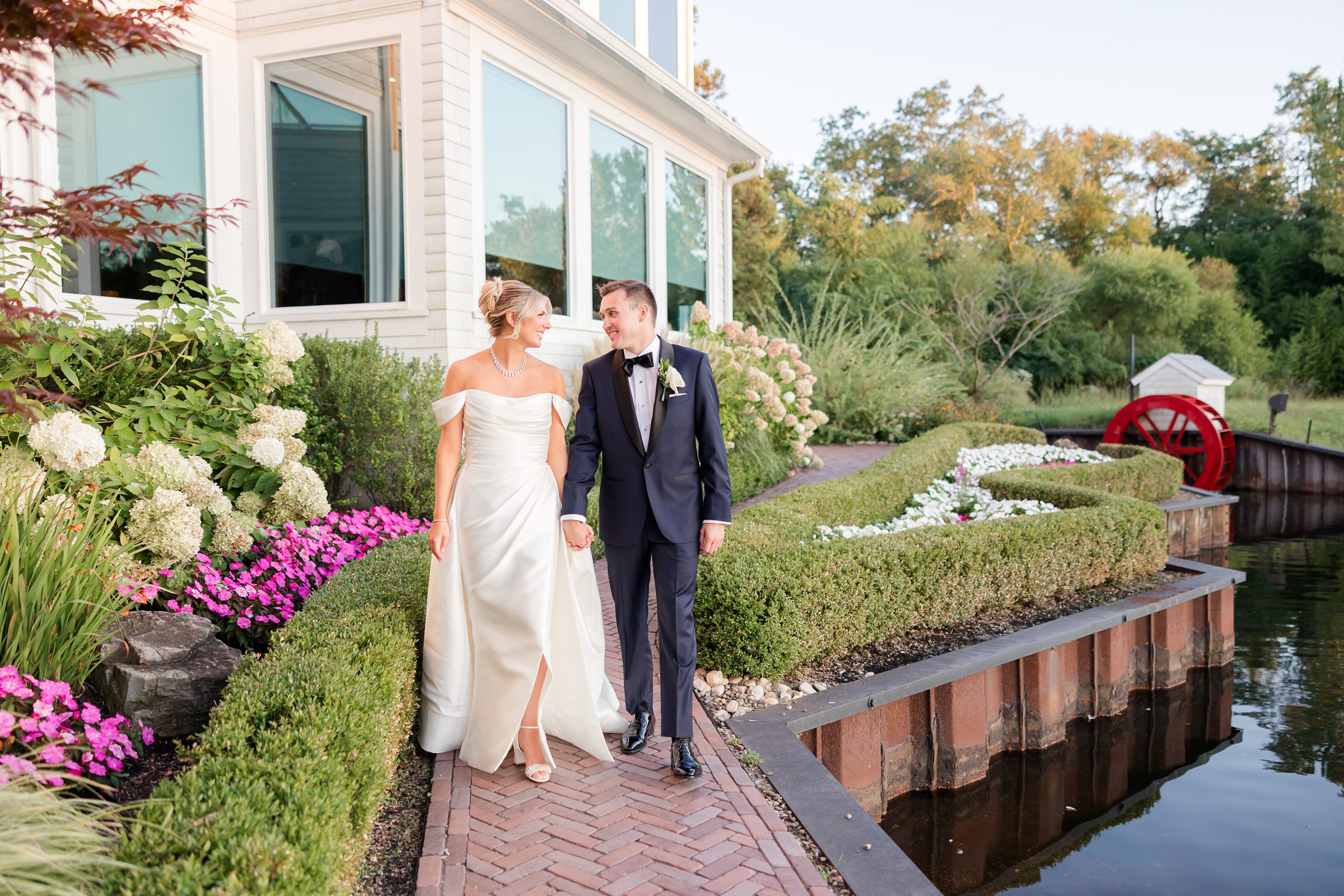 Bride and groom walk together along a brick path beside a waterfront garden, smiling at each other in soft evening light.
