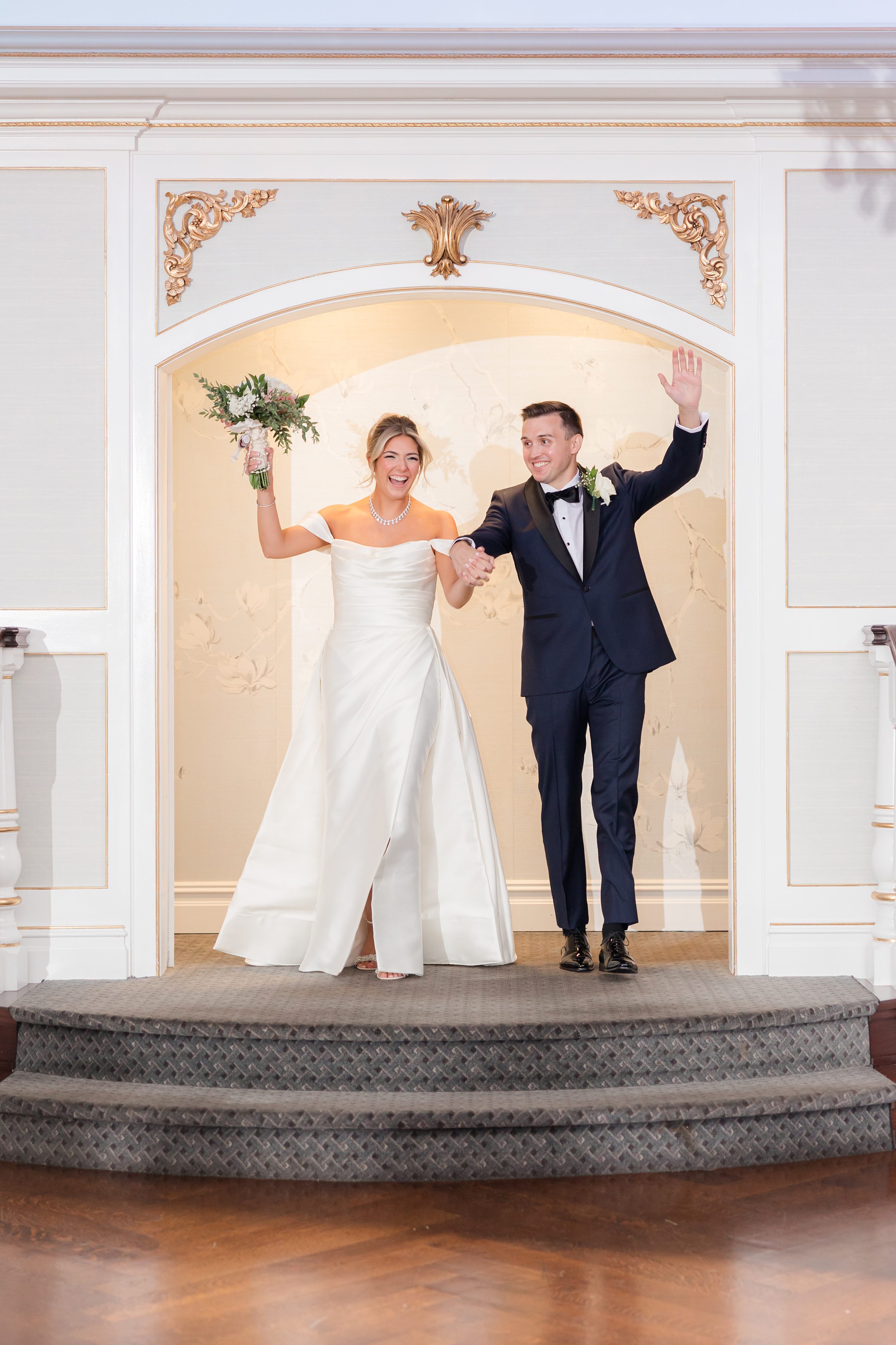 Bride and groom walk together through an elegant doorway, smiling with pure joy as they make their grand entrance, the bride lifting her bouquet and the groom waving in celebration of their new beginning together.