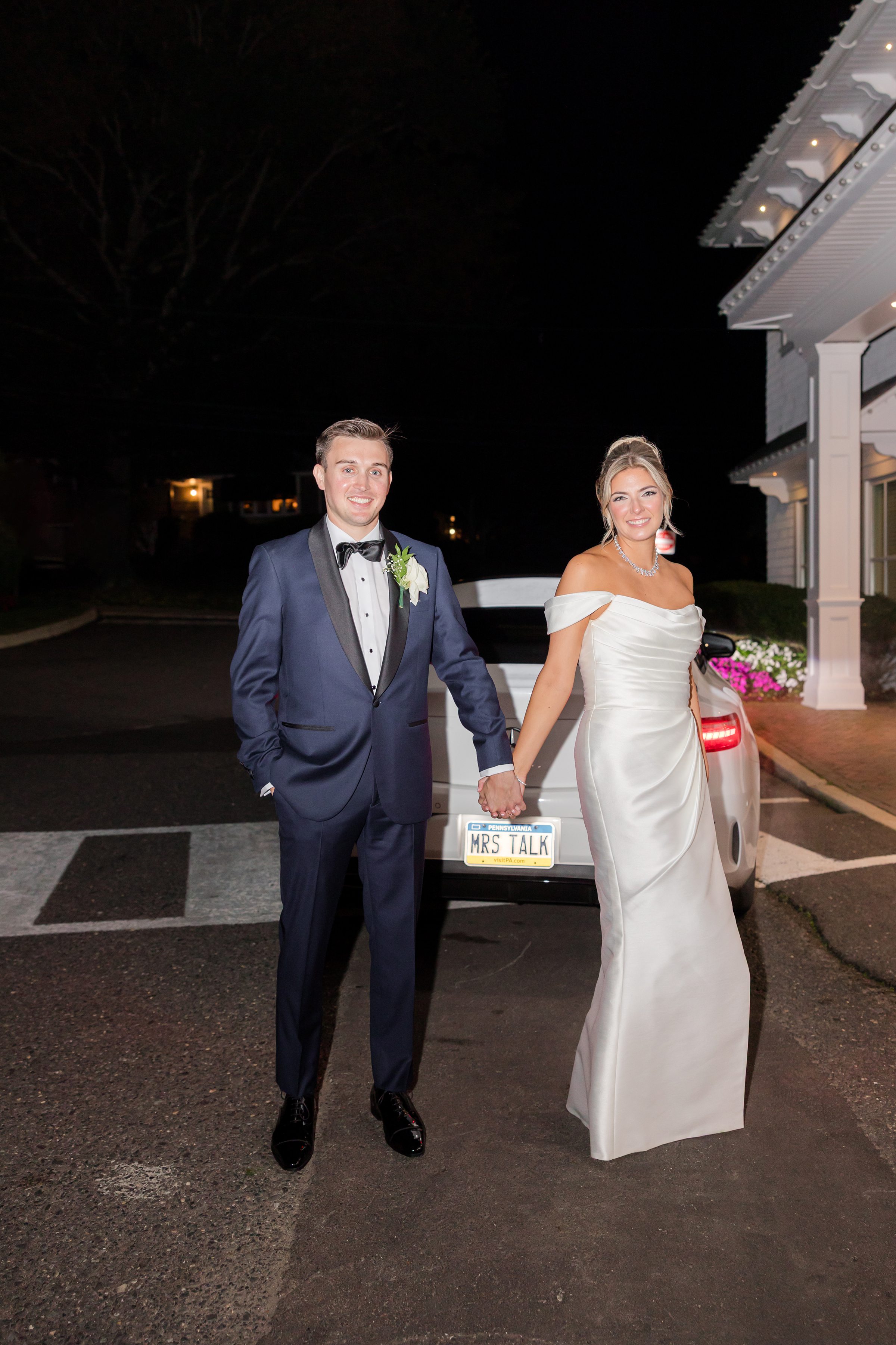 Bride and groom stand together at night, fingers intertwined, smiling softly as they pause in a peaceful moment after their celebration.