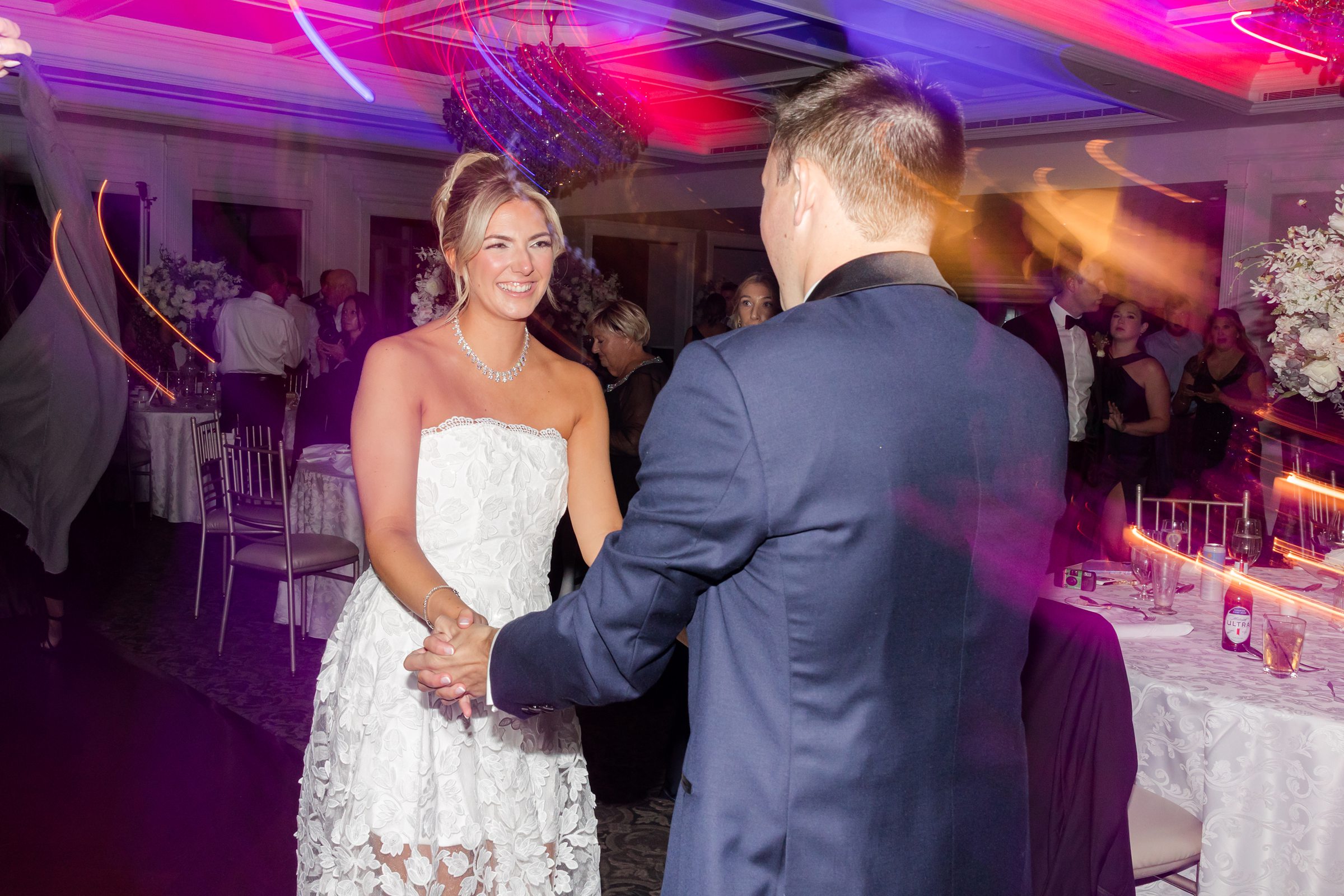 Bride and groom shares a sweet moment on the dance floor, holding hands and smiling as colorful lights swirl around them, capturing the magic of their celebration.