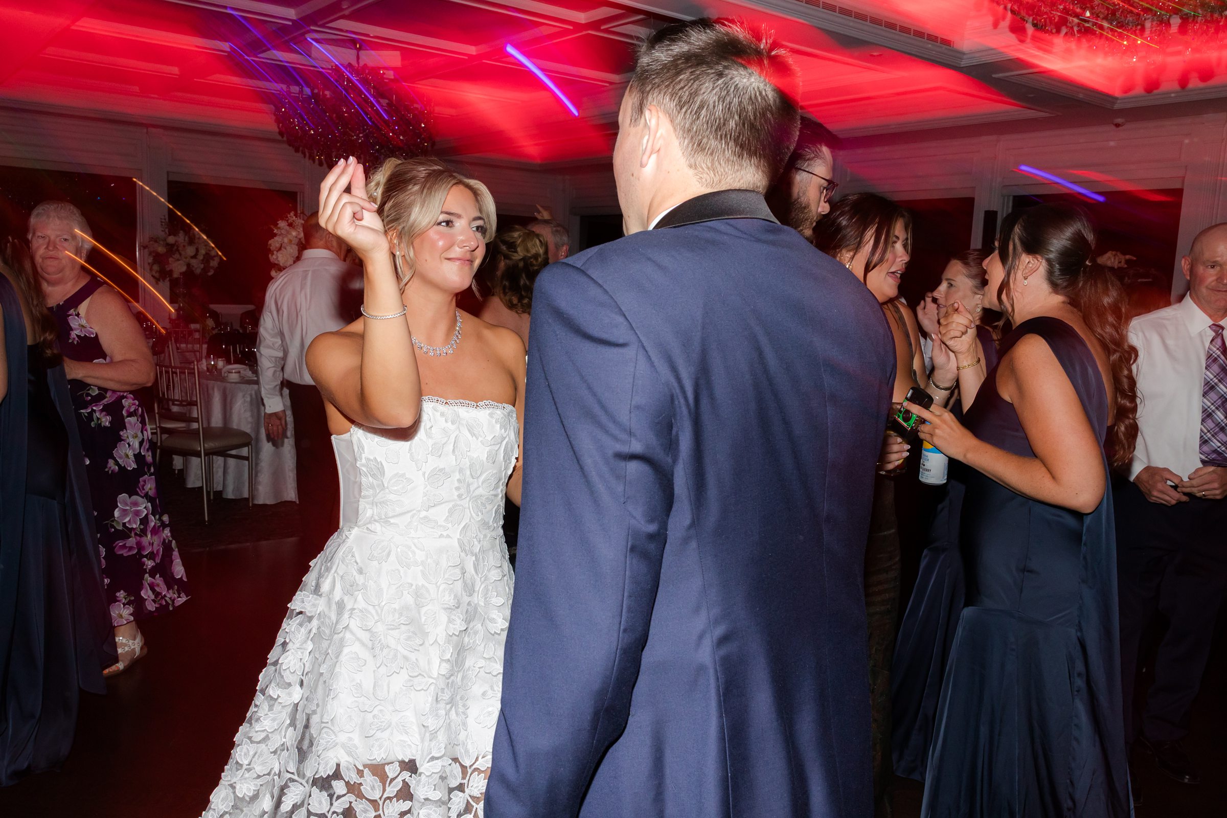 Bride dances playfully under glowing red lights, her expression soft and full of love as she faces her groom across the crowded reception floor.