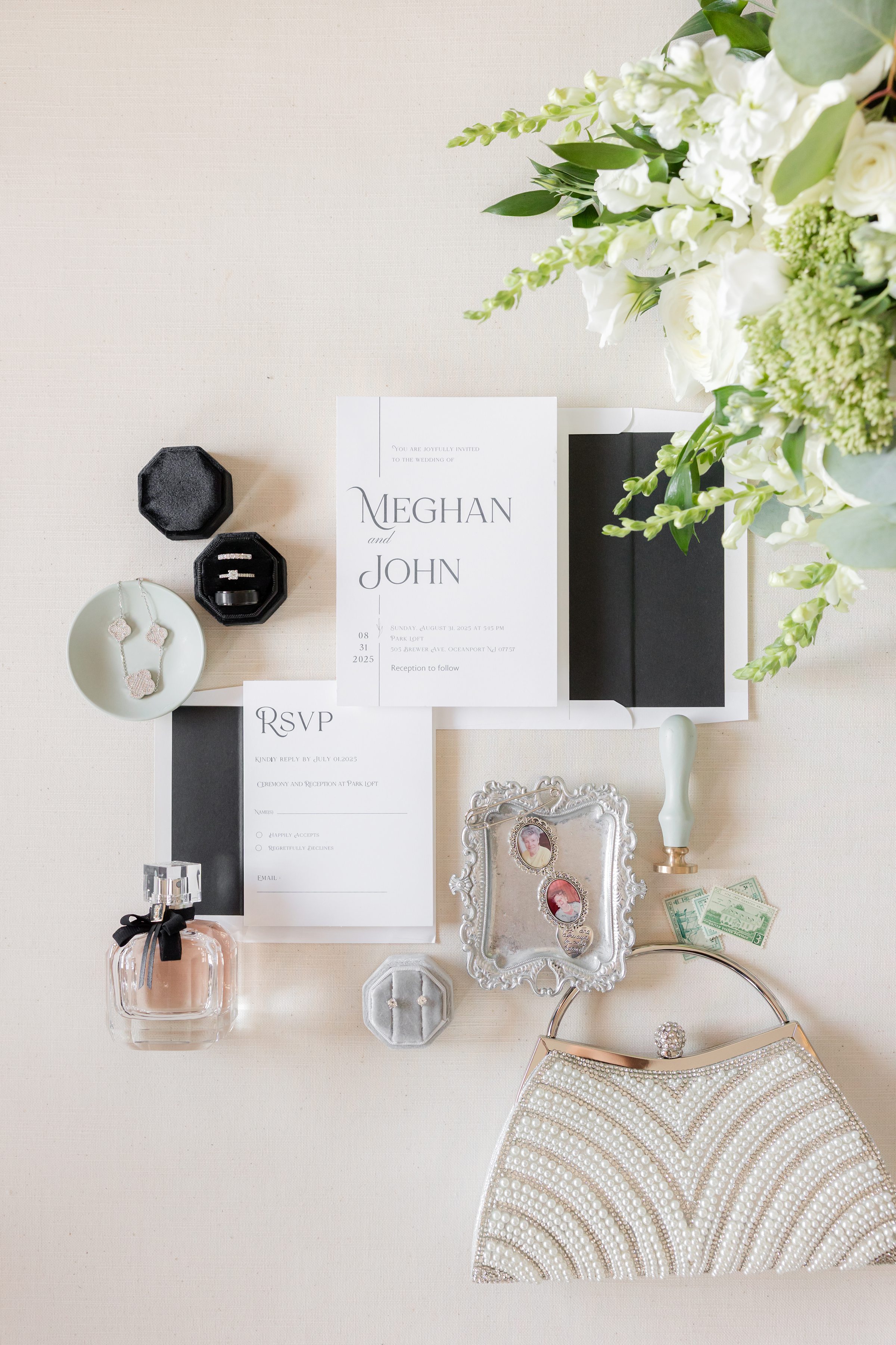 Wedding stationery flat lay featuring a soft ivory invitation suite for Meghan and John, paired with sparkling rings in velvet boxes, a vintage silver heirloom frame, delicate perfume, pastel jewelry, and a beaded bridal clutch—romantically styled to evoke timeless elegance and the anticipation of the wedding day